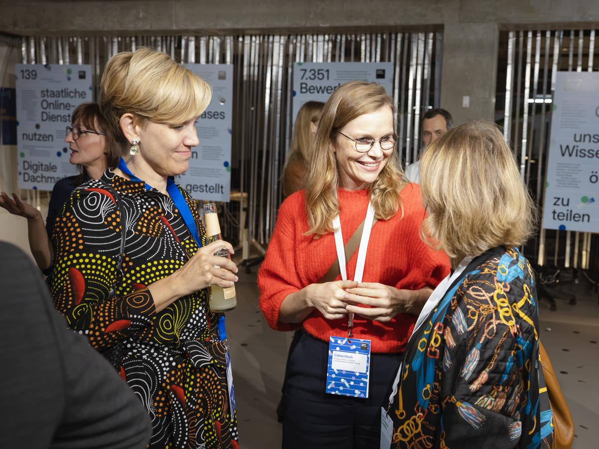 Drei Gästinnen des Events stehen zusammen und unterhalten sich, im Hintergrund sind Plakete mit Zahlen zur Arbeit des DigitalService zu sehen