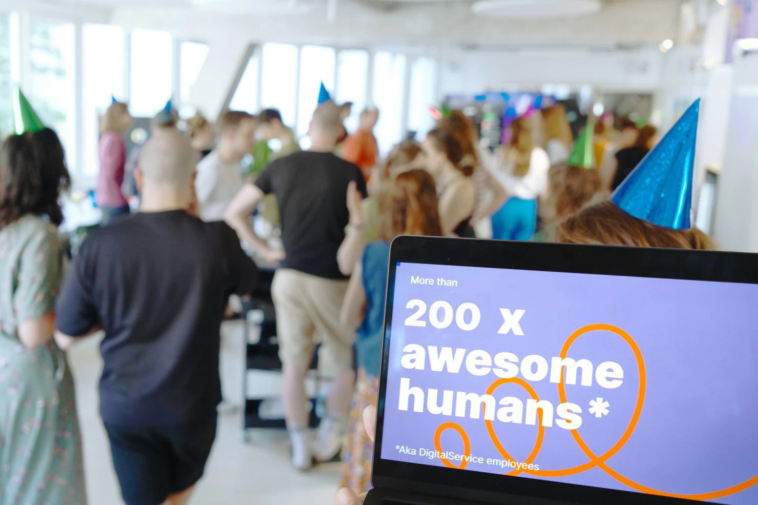 Im Vordergrund ist auf einem Laptopbildschirm zu lesen, dass mehr als 200 Mitarbeitende nun beim DigitalService arbeiten; im Hintergrund sind unscharf zahlreiche Mitarbeitende zu erkennen, die gemeinsam feiern