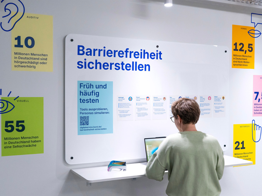 Eine Person sitzt mit einem Laptop an einem Tisch vor einer großen weißen Infotafel mit der Überschrift „Barrierefreiheit sicherstellen“ im Empathie-Labor des DigitalService. Die Tafel zeigt Informationen zu verschiedenen Behinderungen und Statistiken in Deutschland, wie „10 Millionen Menschen in Deutschland sind hörgeschädigt oder schwerhörig“.