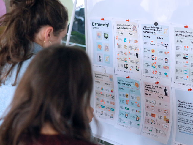 Zwei Personen stehen vor einem Whiteboard an dem verschiedene Zettel mit Hinweisen zur richtigen und falschen Gestaltung von barrierefreien Produkten hängen