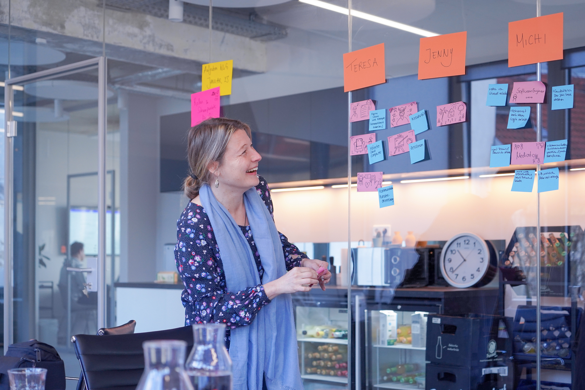 Eine Person steht vor einer Glaswand und klebt dort bunte Zettel an. Auf den größten und am höchten hängenden Zetteln stehen Namen.