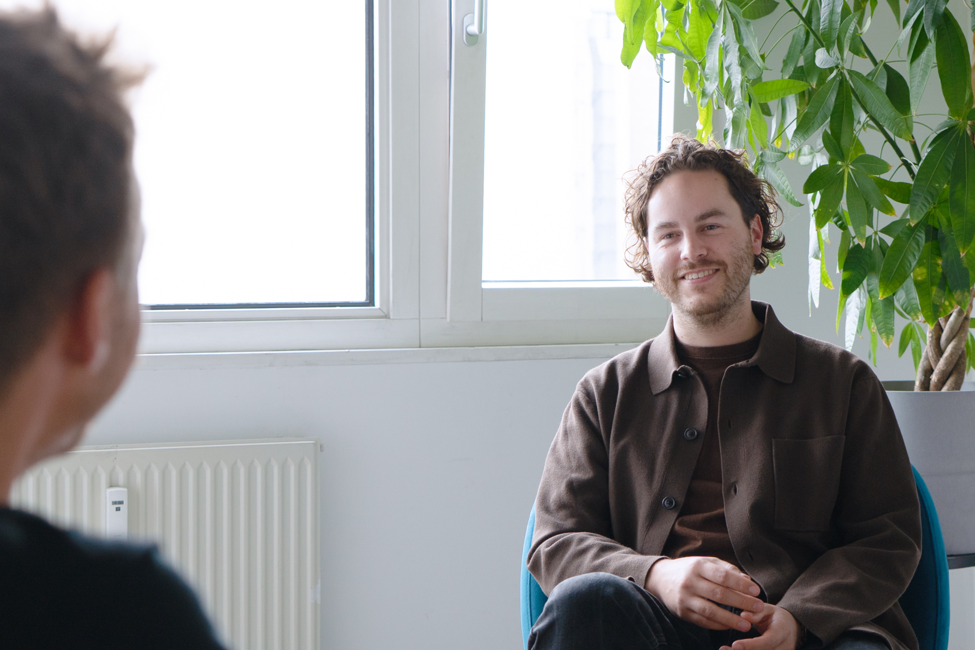 Joshua sitzt in einem modernen Büroraum und lächelt die interviewende Person an. Diese ist undeutlich im Vordergrund zu erkennen. Im Hintergrund fällt Licht durch ein Fenster.