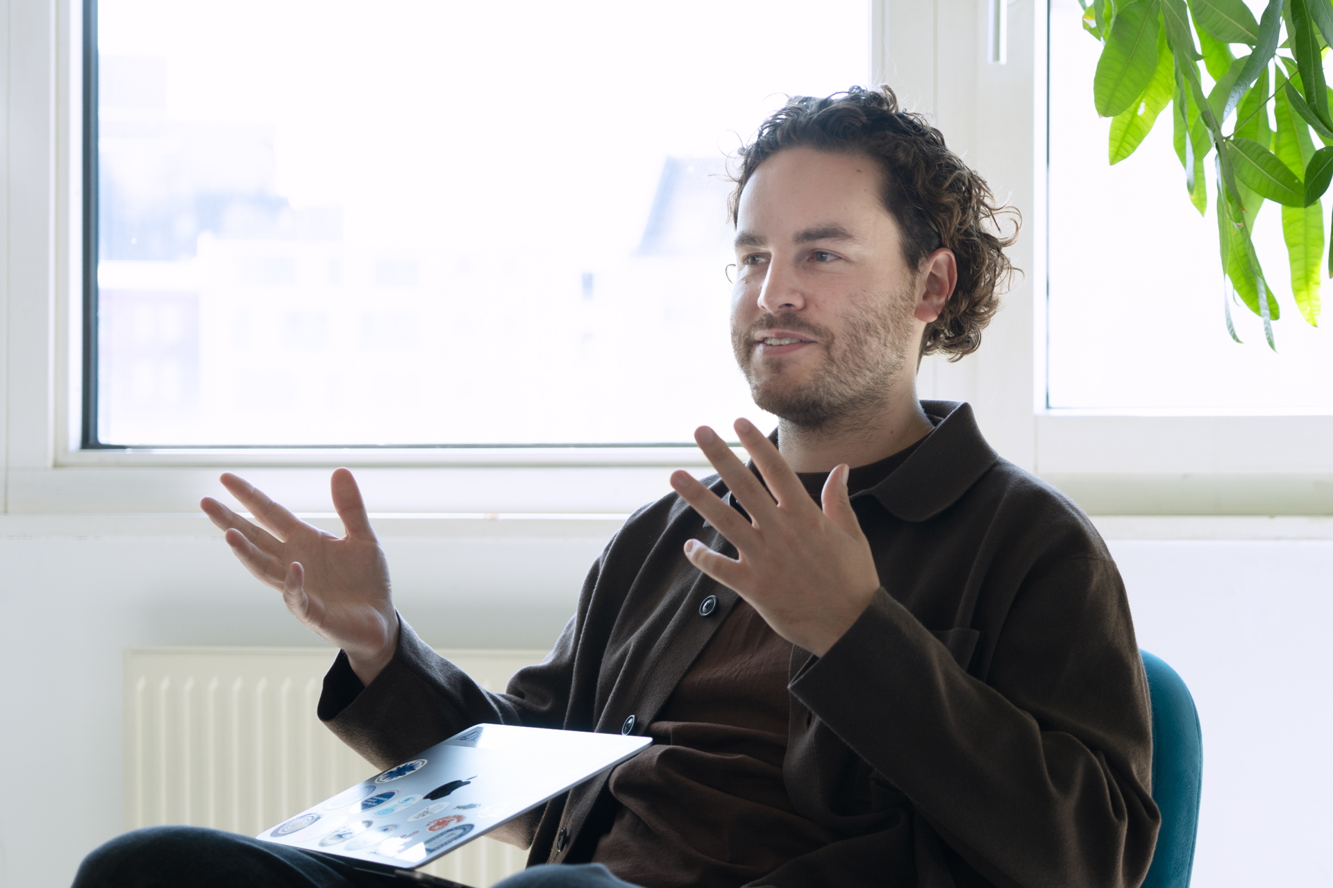 Johsua sitzt in einem modernen Büroraum, gestikuliert mit den Händen und spricht. Auf seinen Beinen steht ein Laptop mit angeklappten Bildschirm, im Hintergrund fällt Licht durch ein Fenster.