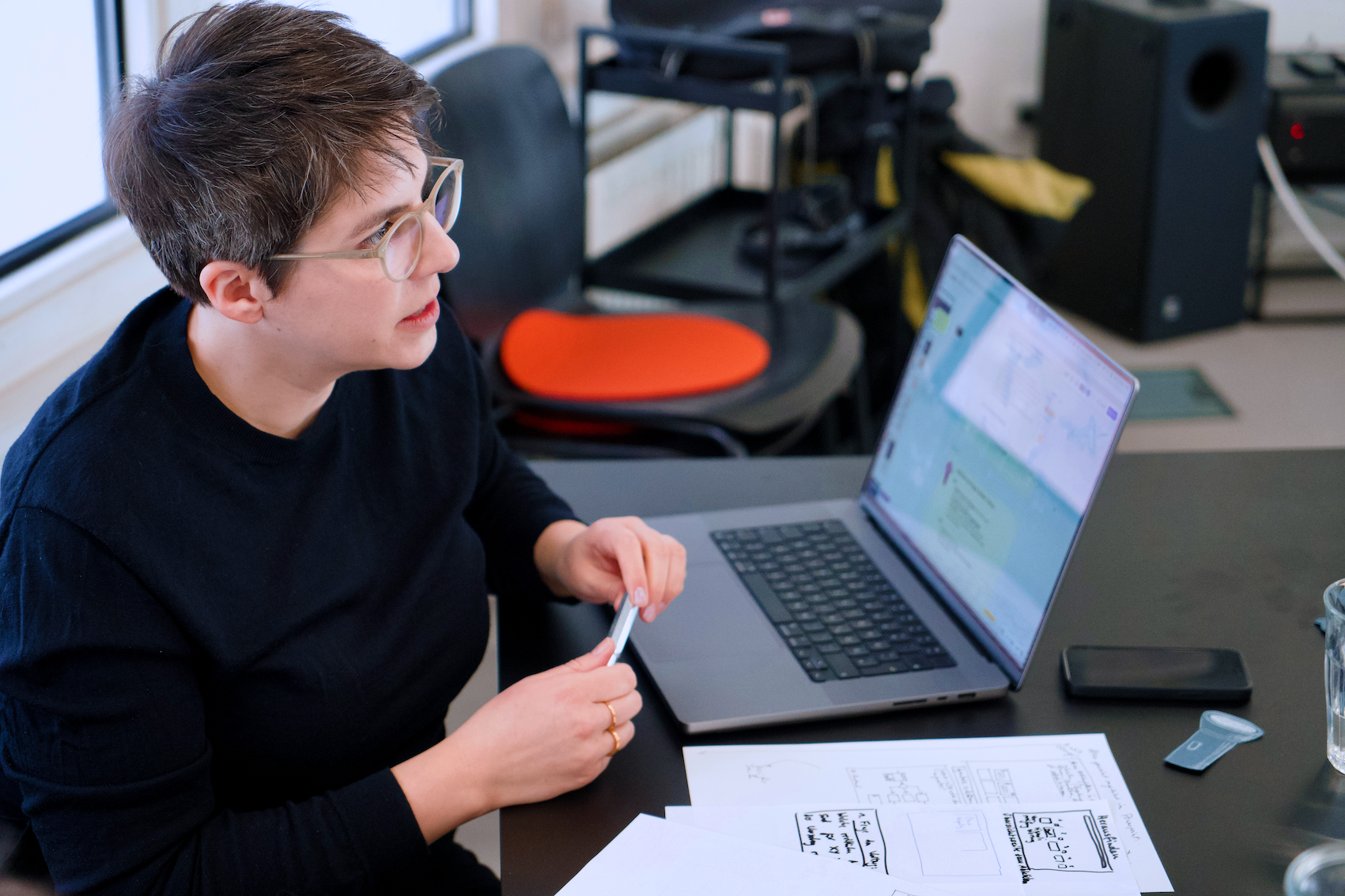 Eine Person sitzt an einem schwarzen Schreibtisch in einem modernen Büroraum hält einen Stift in der Hand und redet. Vor der Person steht ein Laptop. Auf dem geöffneten Bildschirm läuft ein Programm. Auf dem Tisch liegen Notizen und ein Handy. Im Hintergrund sind Bürogegenstände zu sehen.