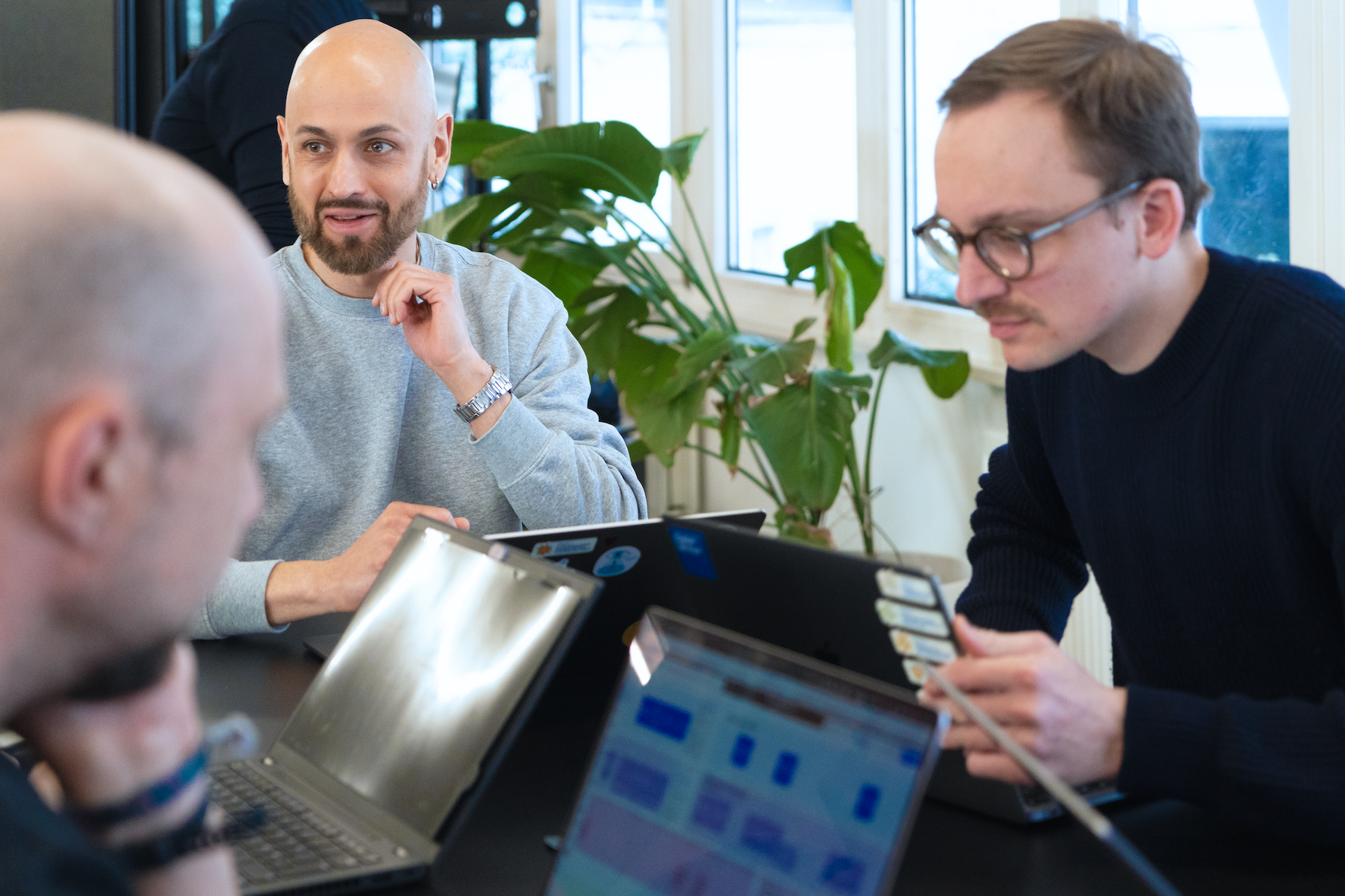 Mehrere Personen sitzen an einem schwarzen Schreibtisch, reden und schauen auf ihre geöffneten Laptops. Im Hintergrund ist eine moderne Büroumgebung mit einer Fensterfront und einer grünen Pflanze.