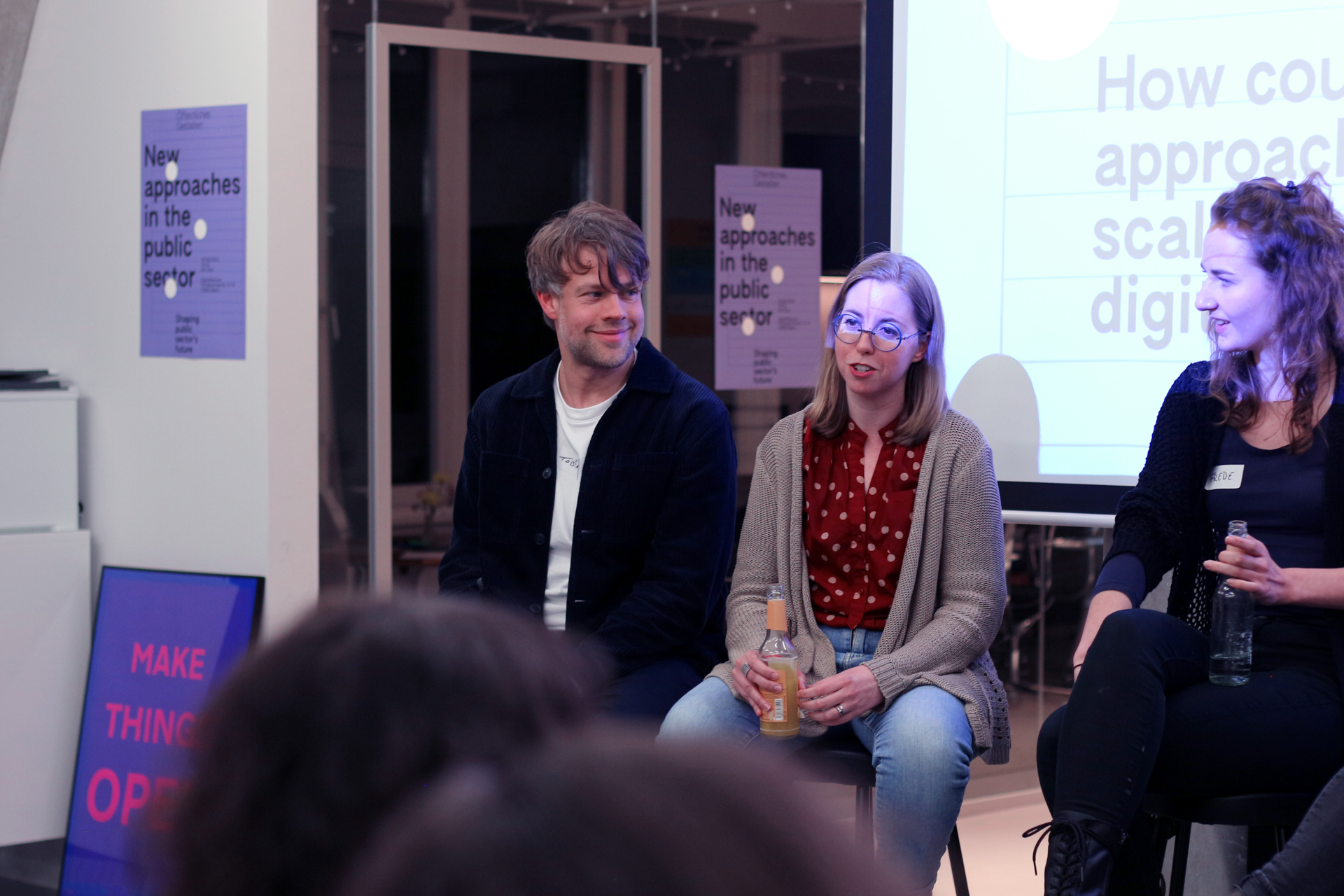 Three people, two women and one man, with lighter skin tones sit in front of a screen and audience and look at each other - around them hang posters that read in English: 