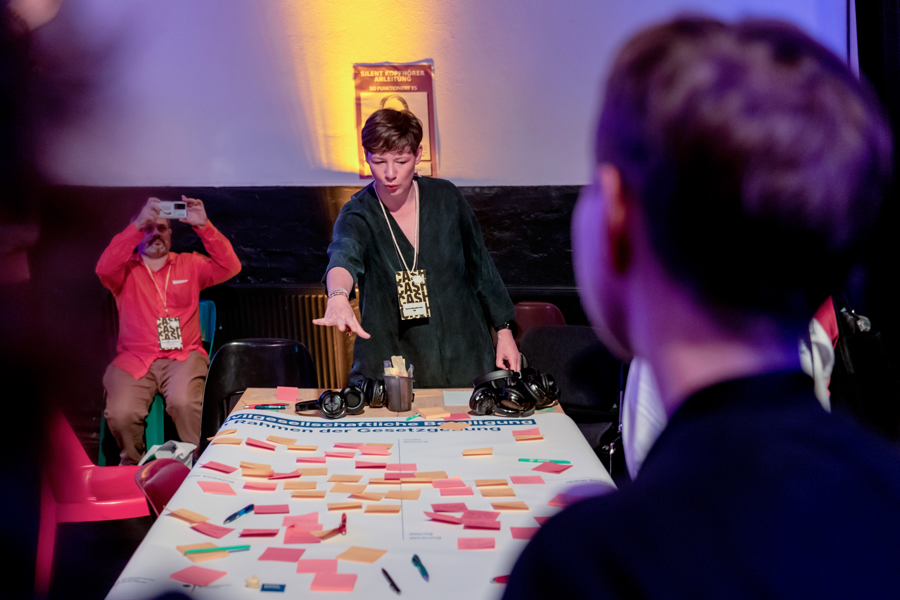 On a table is a larger poster with many notes on colorful post-its. Some people are grouped around it, one person speaks and points to an area of the poster.