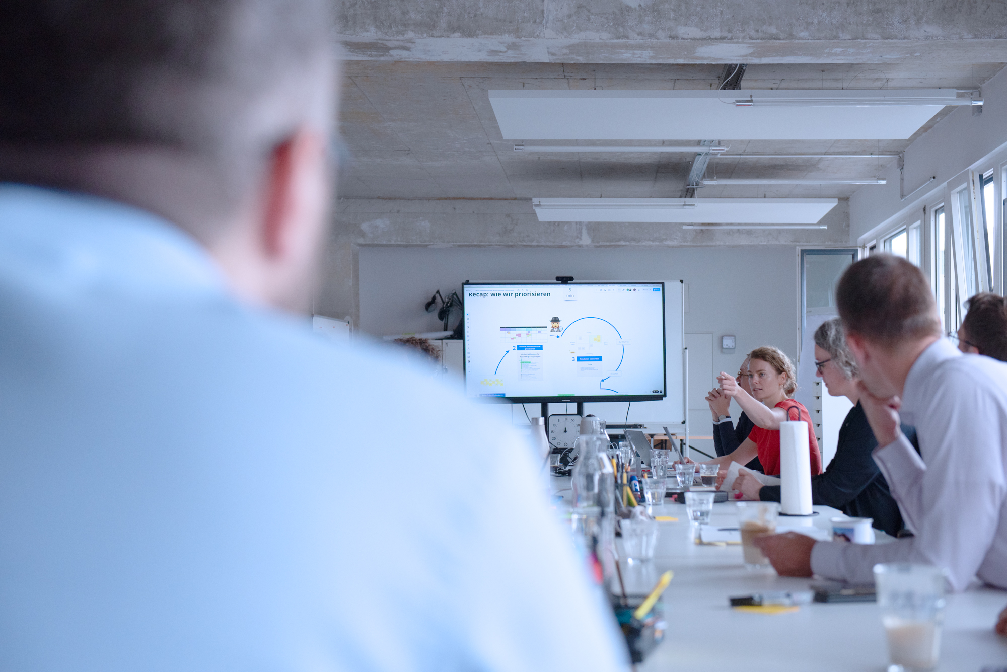 A larger group sits around a conference table and discusses; at the front end, a screen shows a presentation on the step-by-step development of the digital check.