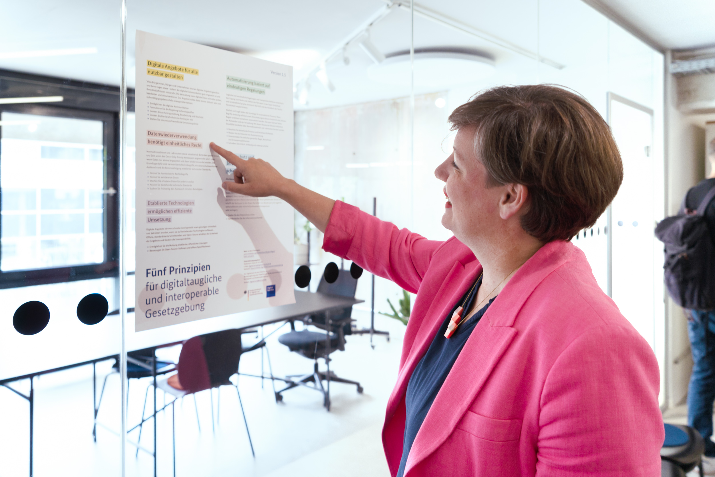 Katrin Lütkemöller Shaw steht in einem modernen Büro und zeigt lächelnd auf ein Poster mit dem Titel „Fünf Prinzipien für digitaltaugliche und interoperable Gesetzgebung“, das an einer Glasscheibe befestigt ist.