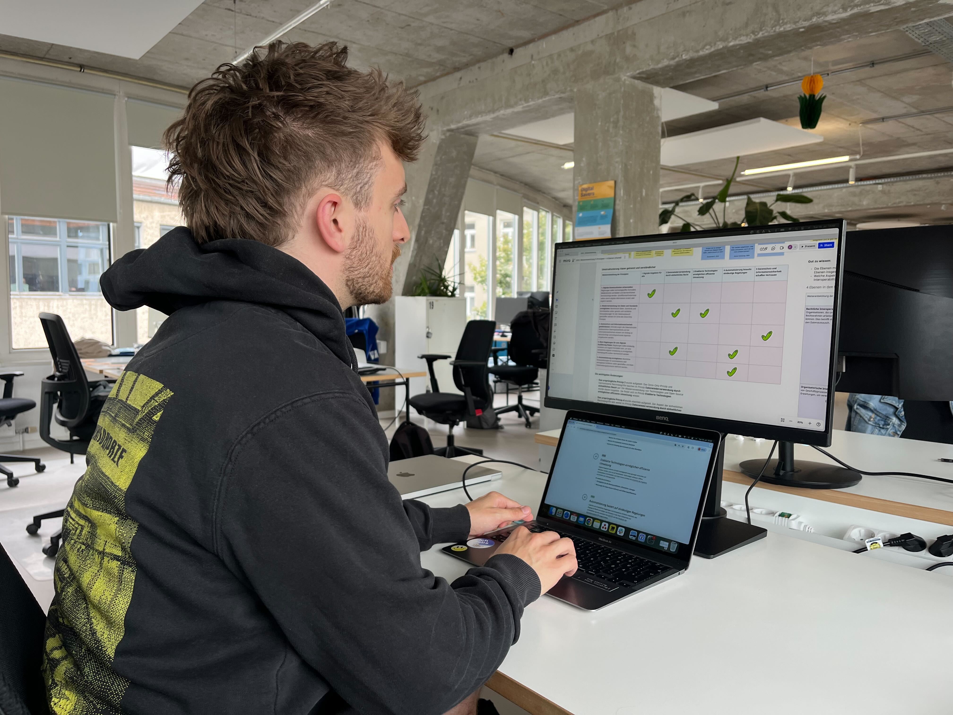 Hendrik Schmidt sits at a desk working on a laptop connected to a large external monitor. The monitor displays a table showing the old and new principles for digitally compatible law.
