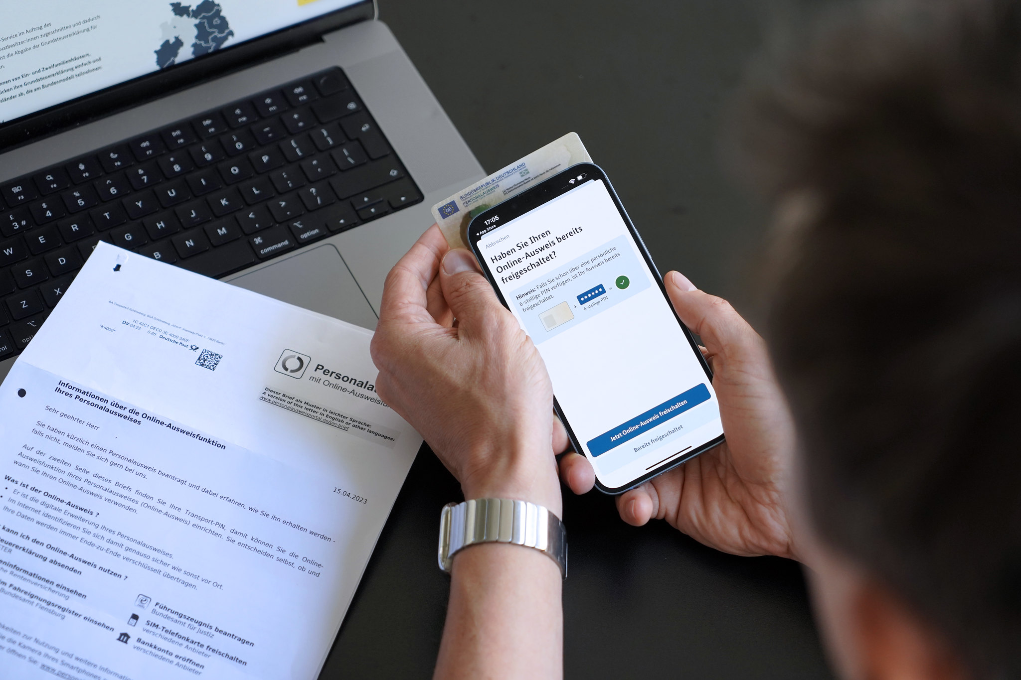 Ein Mann, der ein Smartphone an seinen Personalausweis hält, sitzt an einem Tisch mit einem Behördenschreiben und einem Laptop