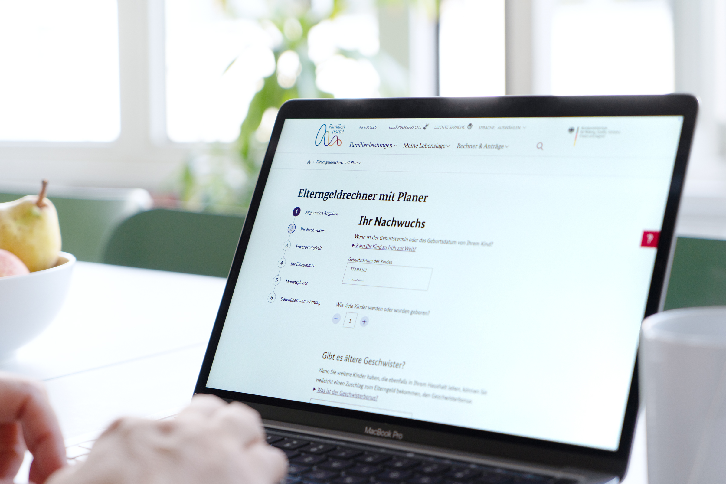 Eine Person sitzt mit einem aufgeklappten Laptop am Tisch und hat die Seite des Elterngeldrechners mit Planer offen