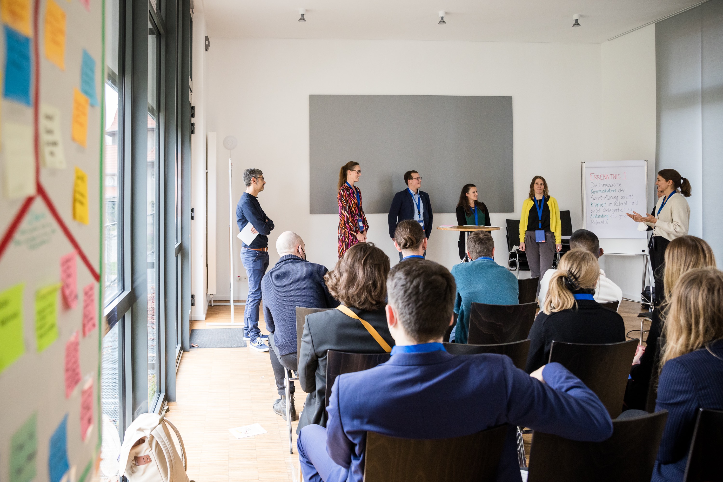 Aufnahme in der Totalen. Im Raum sitzen mindestens 10 Personen im Publikum. Vorne im Raum präsentieren 6 Personen ihre Projektergebnisse. Darunter auch Christian.