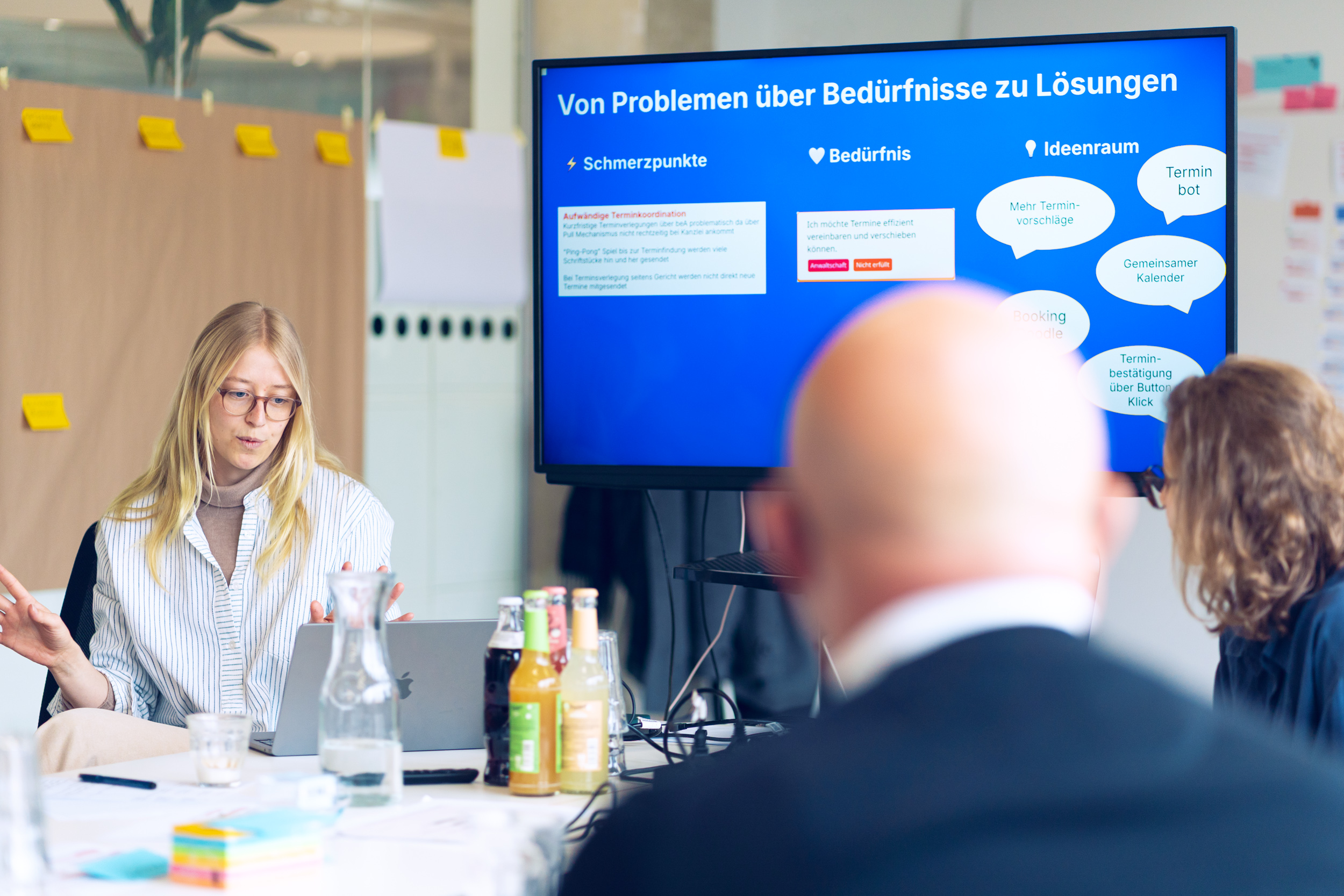 Eine Mitarbeitende des DigitalService präsentiert an einem Laptop, während auf einem Bildschirm hinter ihr eine Folie auf Deutsch mit Überschriften zu Problemen, Bedürfnissen und Lösungen angezeigt wird.