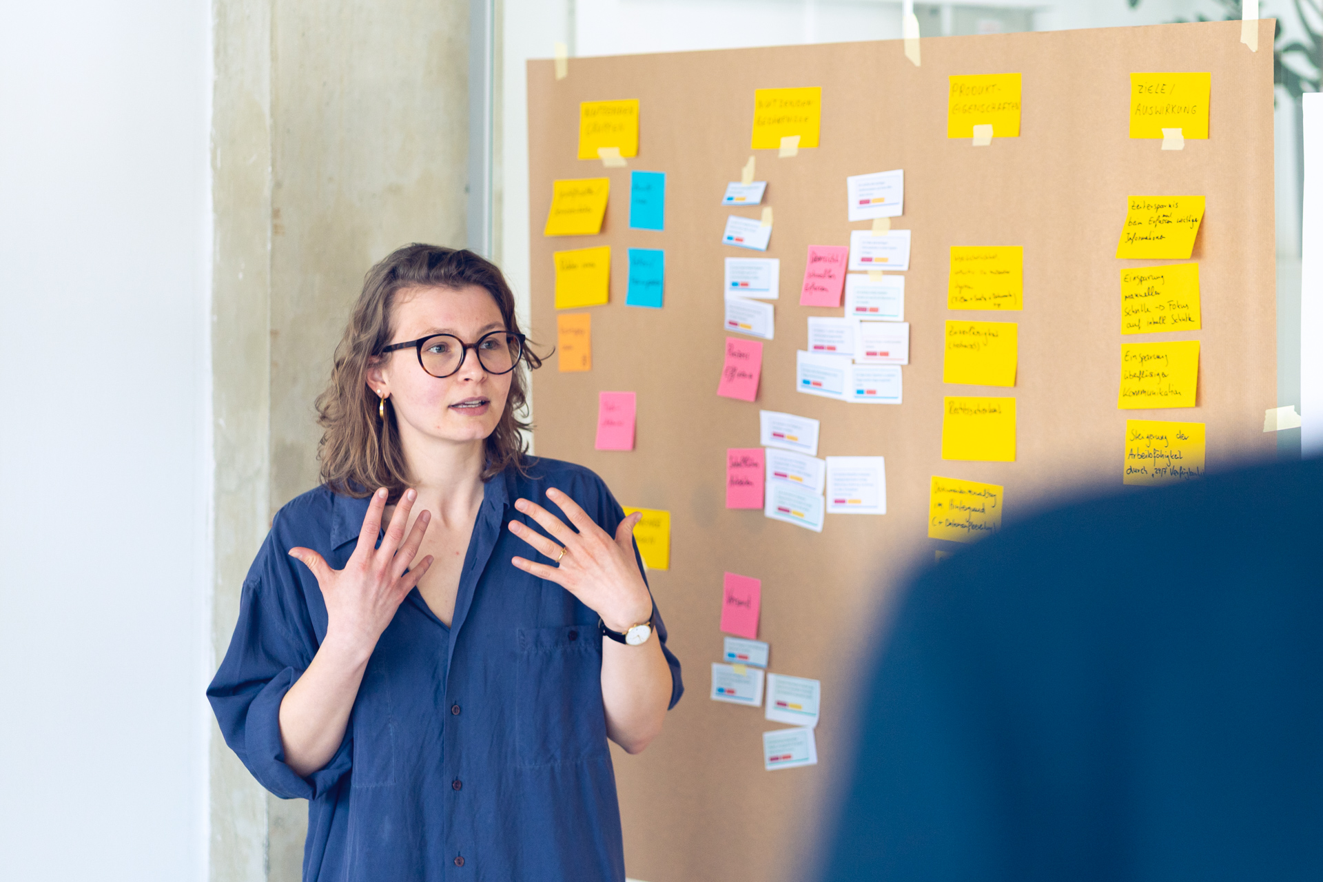 Autorin Lene Baumgart spricht gestikulierend vor einer Wand mit bunten Haftnotizen und Kärtchen, die in Spalten angeordnet sind.