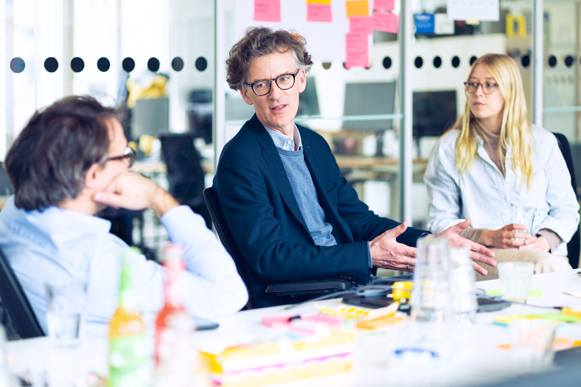 Mitglieder des Projektteams Kommunikationsplattform sitzen an einem Tisch mit Getränken und Büromaterial, eine der Personen spricht, während die anderen zuhören.