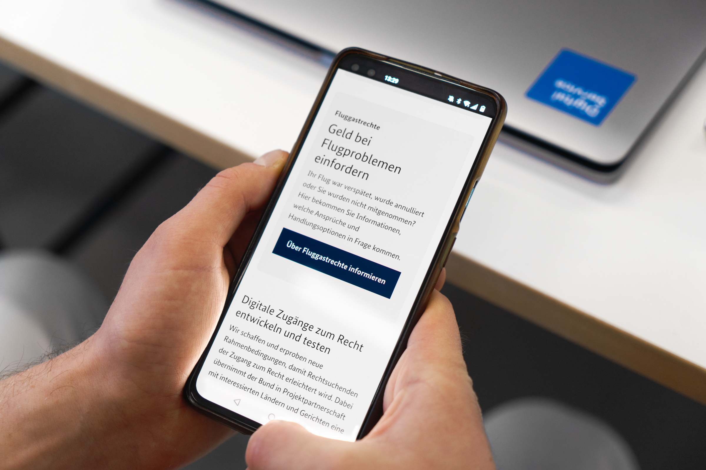 A hand holds a smartphone with a website about air passenger rights. The text shows information on compensation for flight problems. In the background is a laptop with a blue sticker on a table.