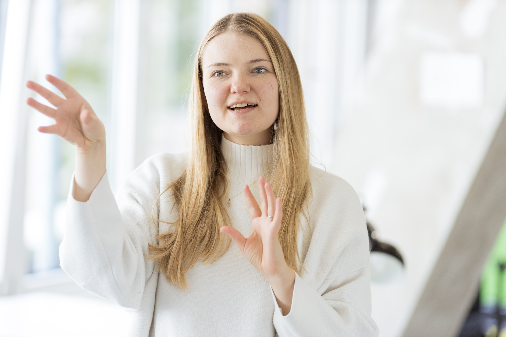 Carolin bewegt ihre Hände, um etwas darzustellen. Die rechte Hand ist dabei höher als die linke Hand und beide Handflächen zeigen nach vorne.