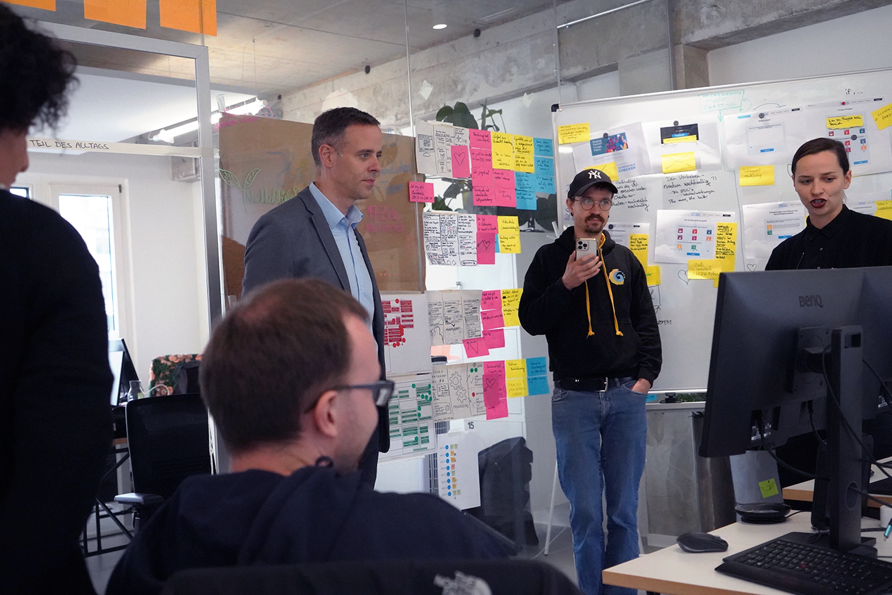 Auf dem Bild stehen mehrere Menchen um einen Bildschirm. CIO Markus Richter bekommt etwas erklärt. Tomie steht in der Mitte des Bildes. Er nutzt eine Software auf seinem Handy um das gesporchene in Textform zu wandeln. Im Hintergrund sind eine Vielzahl von Zetteln zu sehen, diese sind an Wänden aus Glas befästigt.