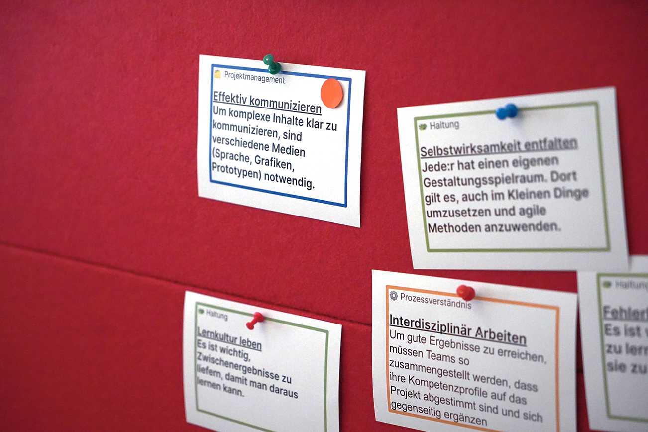 Hier ist eine Rote Wand zu sehen. An der Wand ist eine Vielzahl von Zetteln mit Stecknadeln befästigt. Die Zettel behandeln die verschiedensten ansetze zur effektiven Arbeit in einem Team.