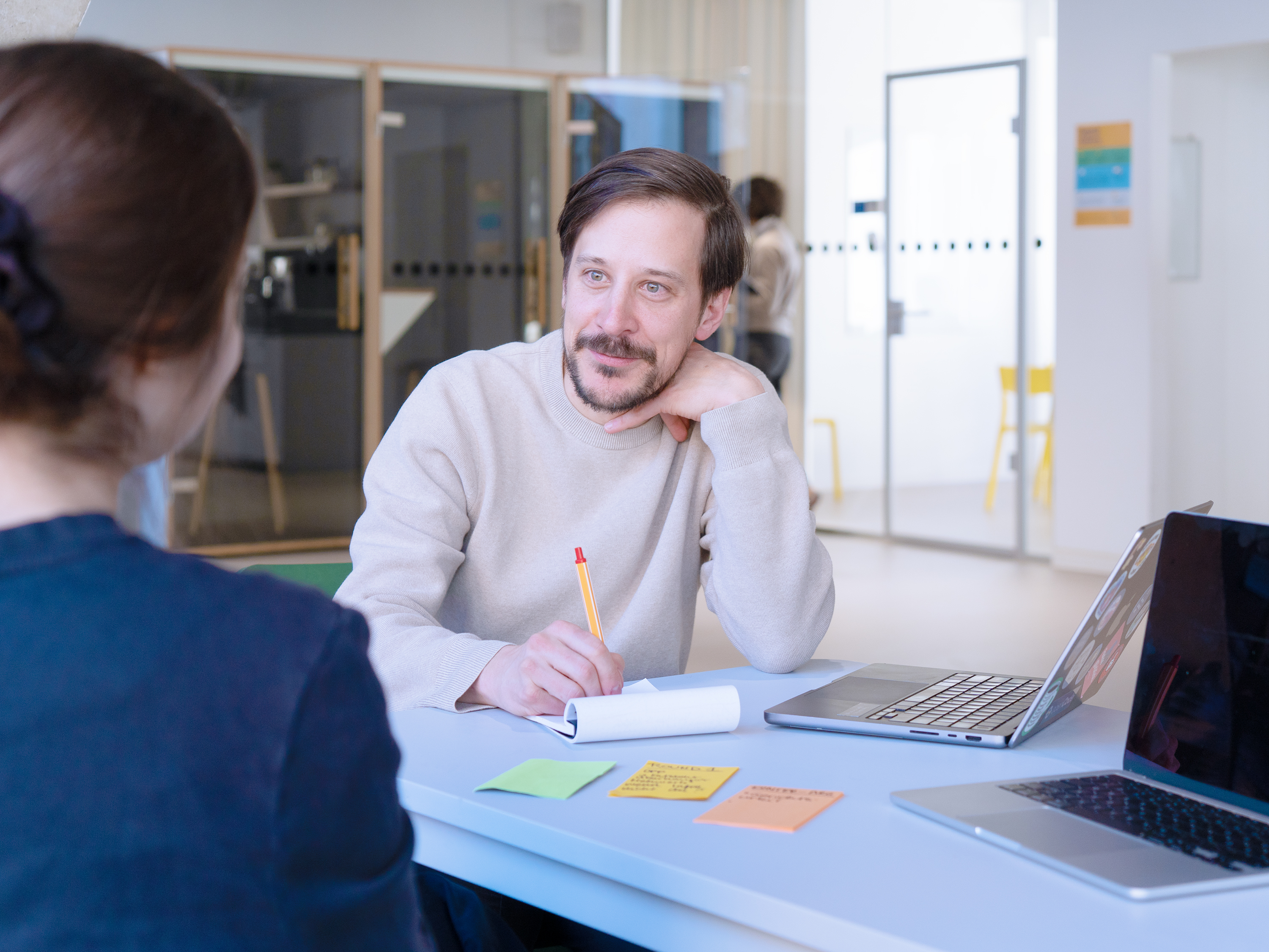 Foto von Niko Felger der in einem modernen Büroraum an einem Schreibtisch sitzt, etwas aufschreibt und Magdalena Zadara interessiert anschaut.