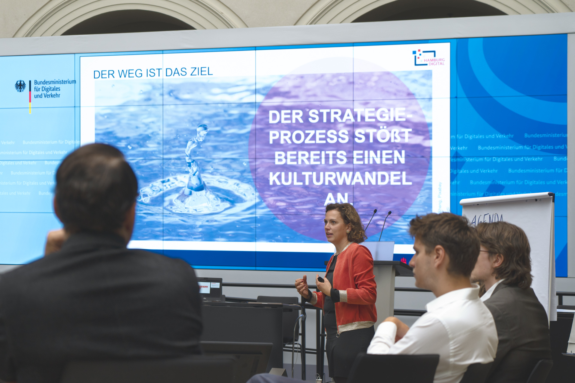 A woman is giving a presentation to a small group. Behind her is a large screen with a picture of a drop of water and the text: “The strategy process is already triggering a cultural change.”