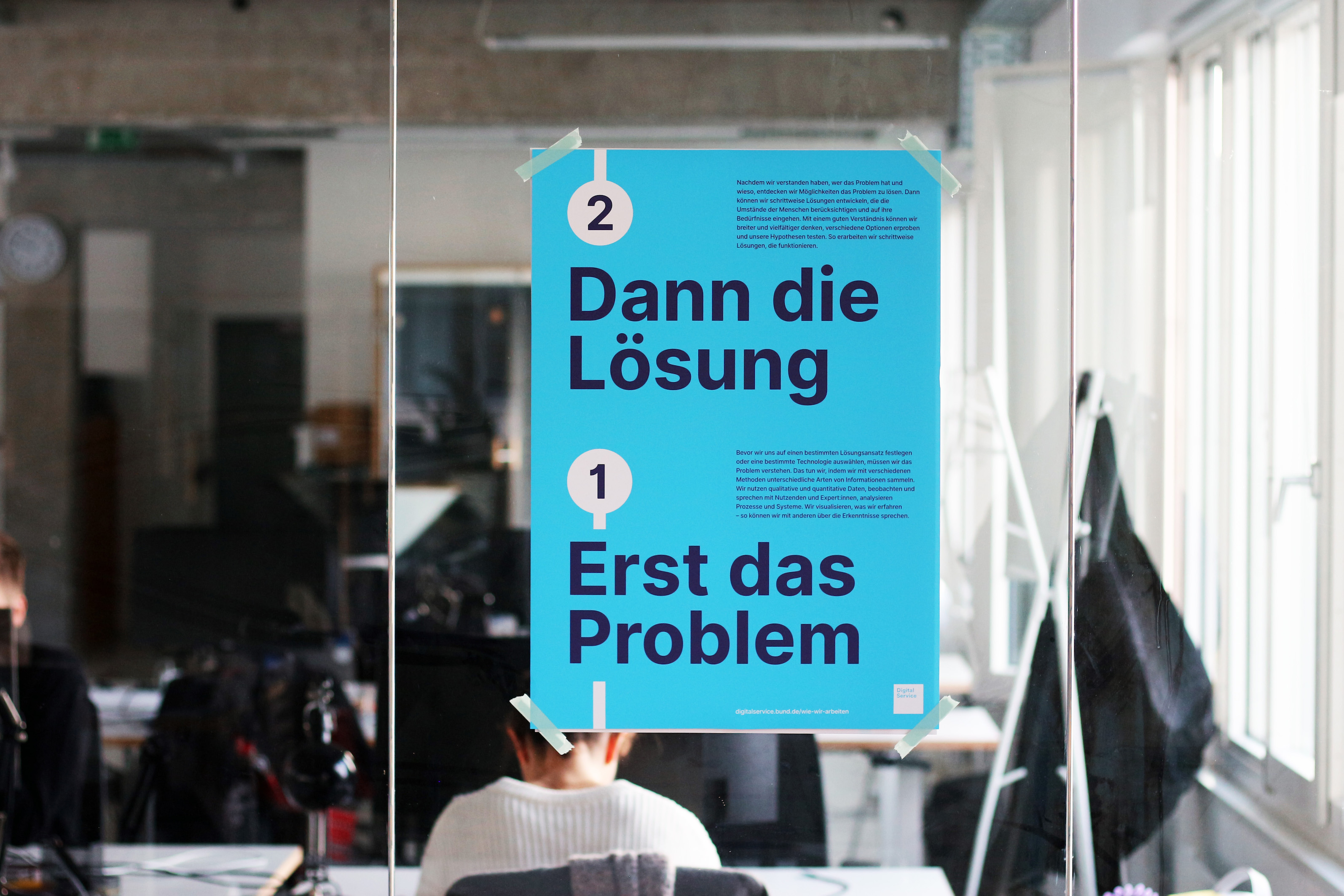 Ein hellblaues Plakat, das an einer Glaswand in einem modernen Büroraum angebracht ist, hinter dem Menschen sitzen. Das Plakat zeigt 2 Schritte im Stil einer U-Bahn-Karte mit Zahlen: 2 Dann die Lösung 1 Erst das Problem Neben den Zahlen ist ein zusätzlicher Text angebracht.