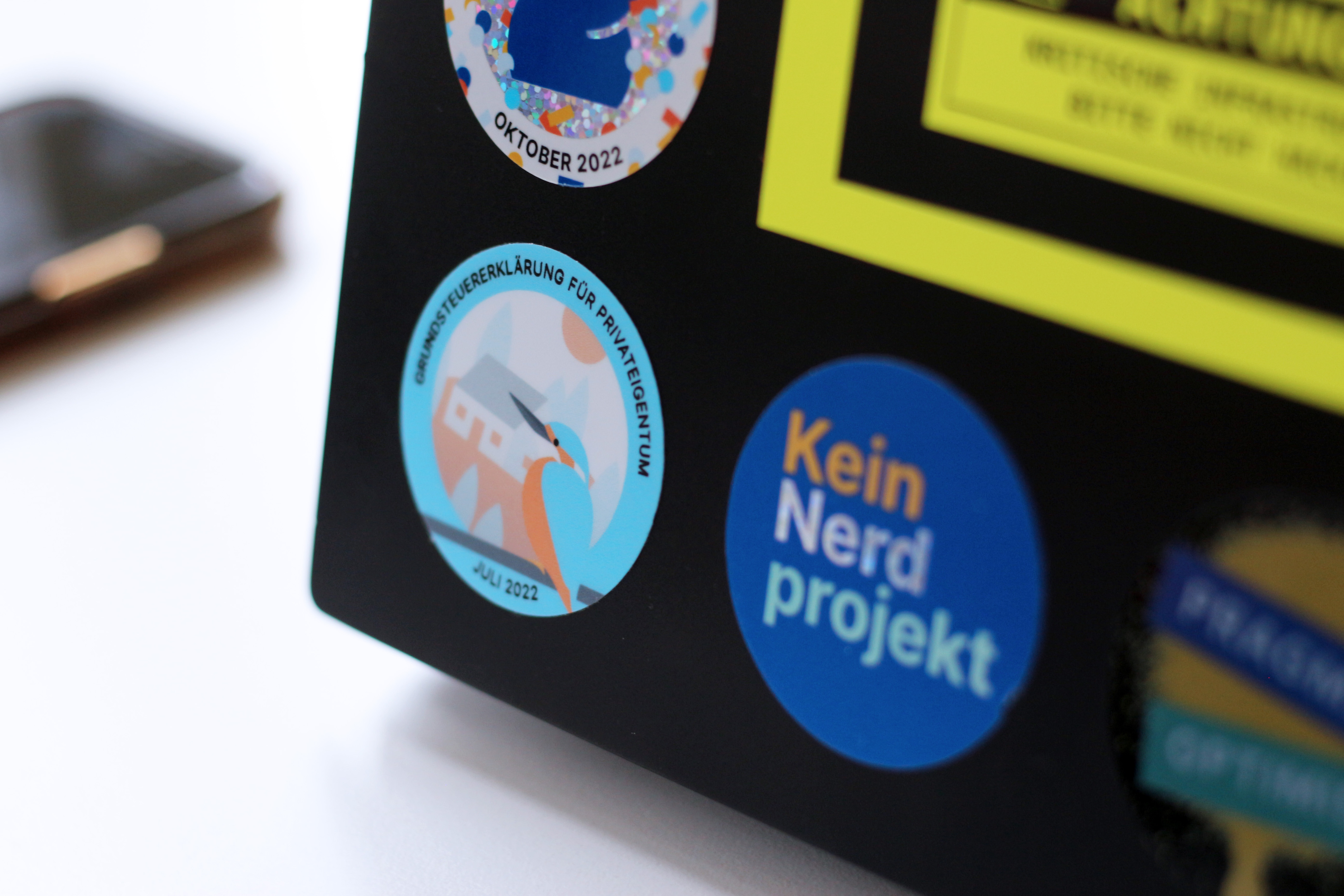 Mehrere bunte Aufkleber auf einem schwarzen Laptop, der auf einem weißen Schreibtisch neben einem Mobiltelefon steht – der Aufkleber im Fokus zeigt einen rot-blauen Vogel vor einem Haus sitzend und dem Aufdruck: „Grundsteuererklärung für Privateigentum – Juli 2022“; ein zweiter Sticker daneben sagt: „Kein Nerdprojekt“, die anderen Aufkleber sind unscharf und angeschnitten