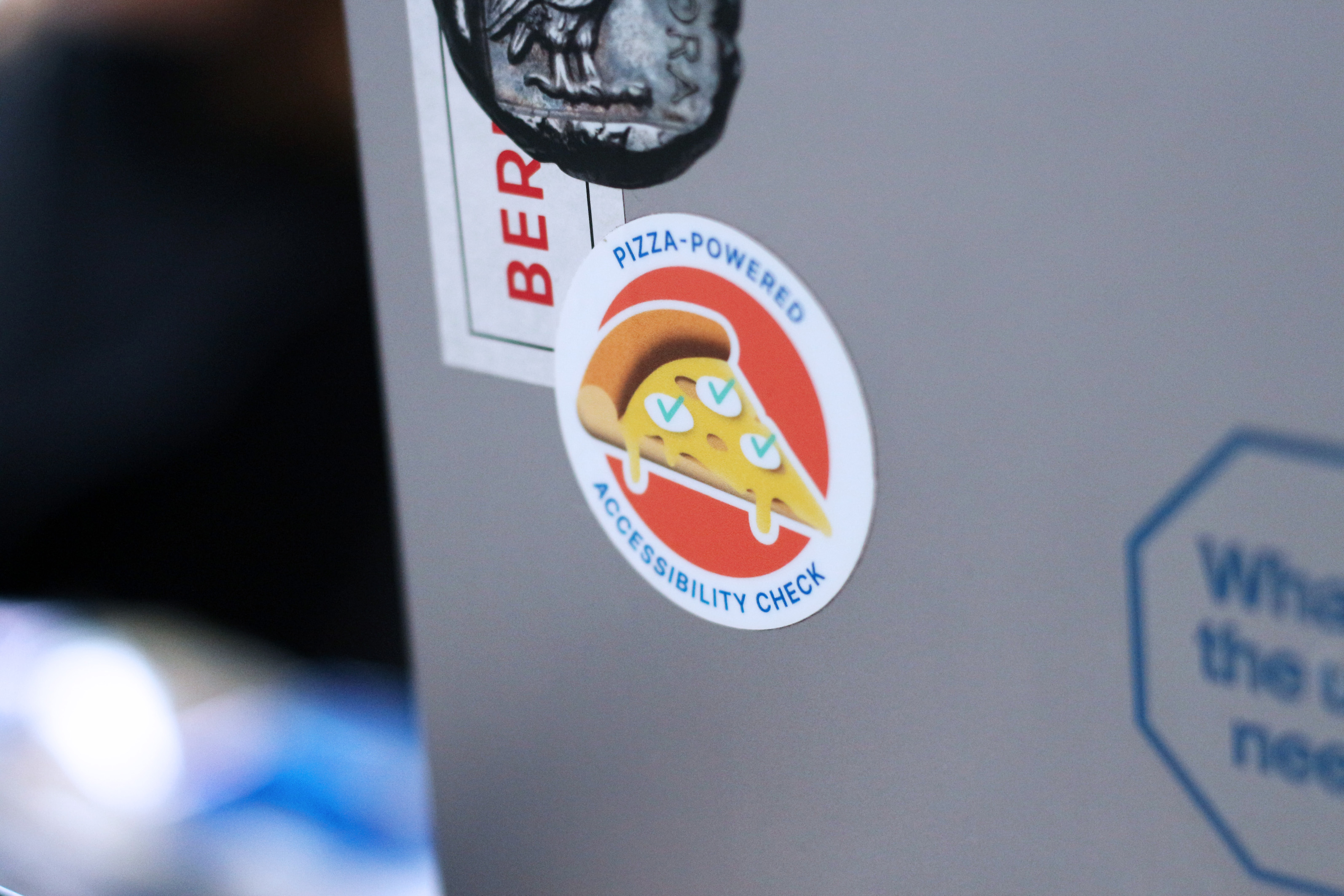 Mehrere Sticker auf einem silbernen Laptop-Computer – der Aufkleber im Fokus zeigt ein Stück Pizza mit geschmolzenem Käse und kleinen grünen Kräutern, die wie kleine grüne Haken ausschauen – drum herum steht in englischer Sprache geschrieben: von Pizza angetriebene Prüfung auf Barrierefreiheit.