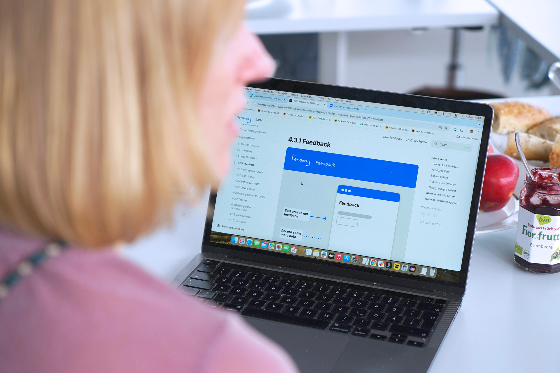 Eine Frau mit blonden Haaren vor einem Laptop-Computer, der die Dokumentation einer Feedback-Komponente von GovStack zeigt; in Hintergrund steht ein Glas Marmelade, Obst und Backwaren