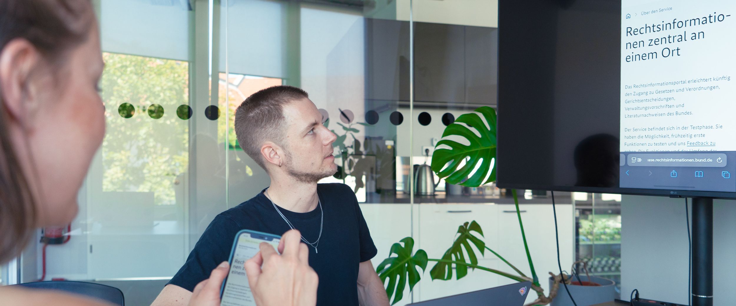 Zwei Personen sitzen in einem modernen Büroraum am Tisch; vor Ihnen steht ein großer Bildschirm mit der Aufschrift „Rechtsinformationen zentral an einem Ort.“ Eine Person sucht Rechtsinformationen auf ihrem Handy; die andere Person lässte diese über ihren Laptop auf dem Bildschirm anzeigen.