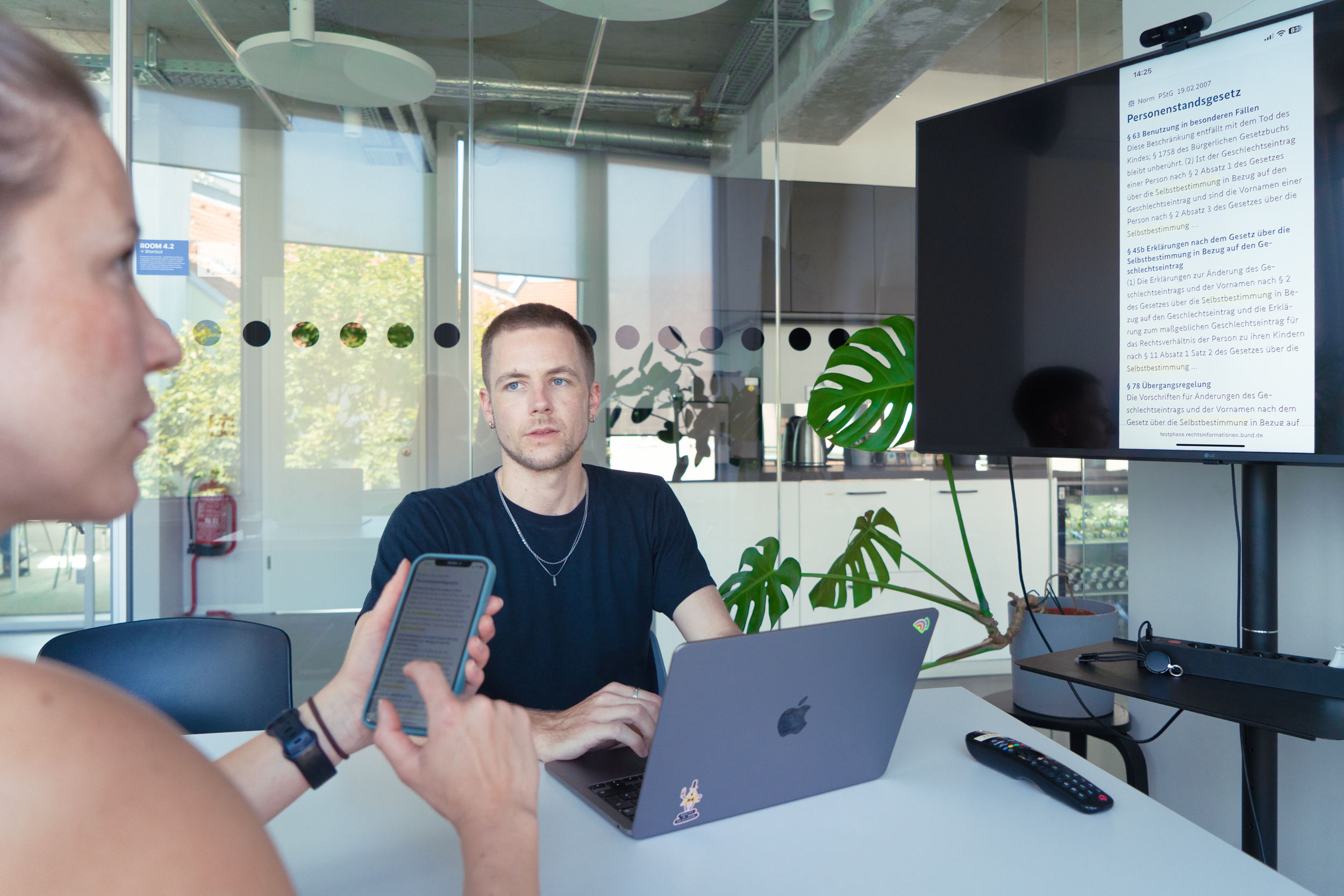 Zwei Personen sitzen in einem modernen Büroraum am Tisch; vor Ihnen steht ein großer Bildschirm mit der Aufschrift „Personenstandgesetz“ Eine Person sucht Rechtsinformationen auf ihrem Handy; die andere Person lässte diese über ihren Laptop auf dem Bildschirm anzeigen.