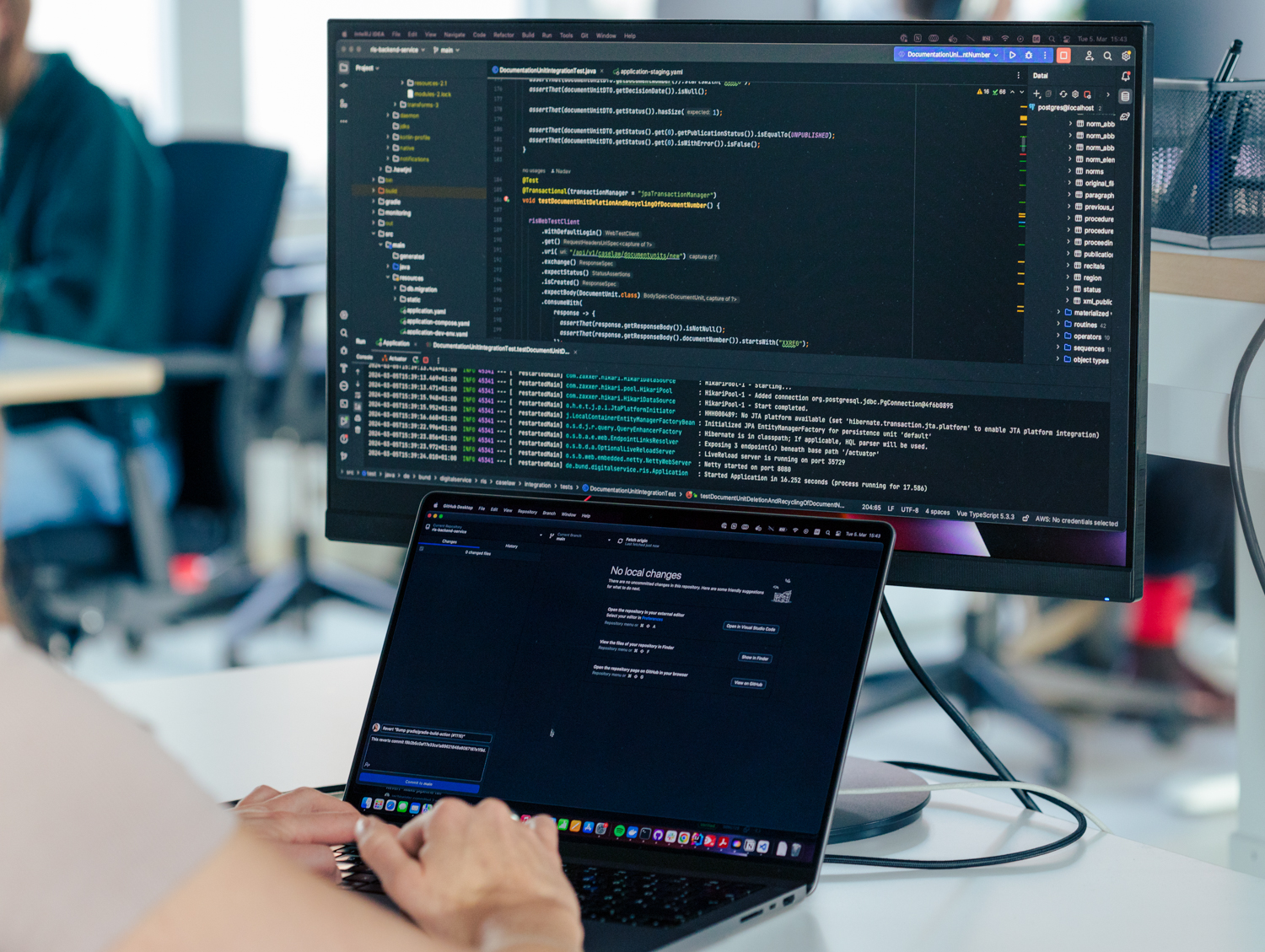 Eine Person sitzt vor einem geöffnente Laptop, dahinter zeigt ein großer Bildschirm Code-Zeilen an.