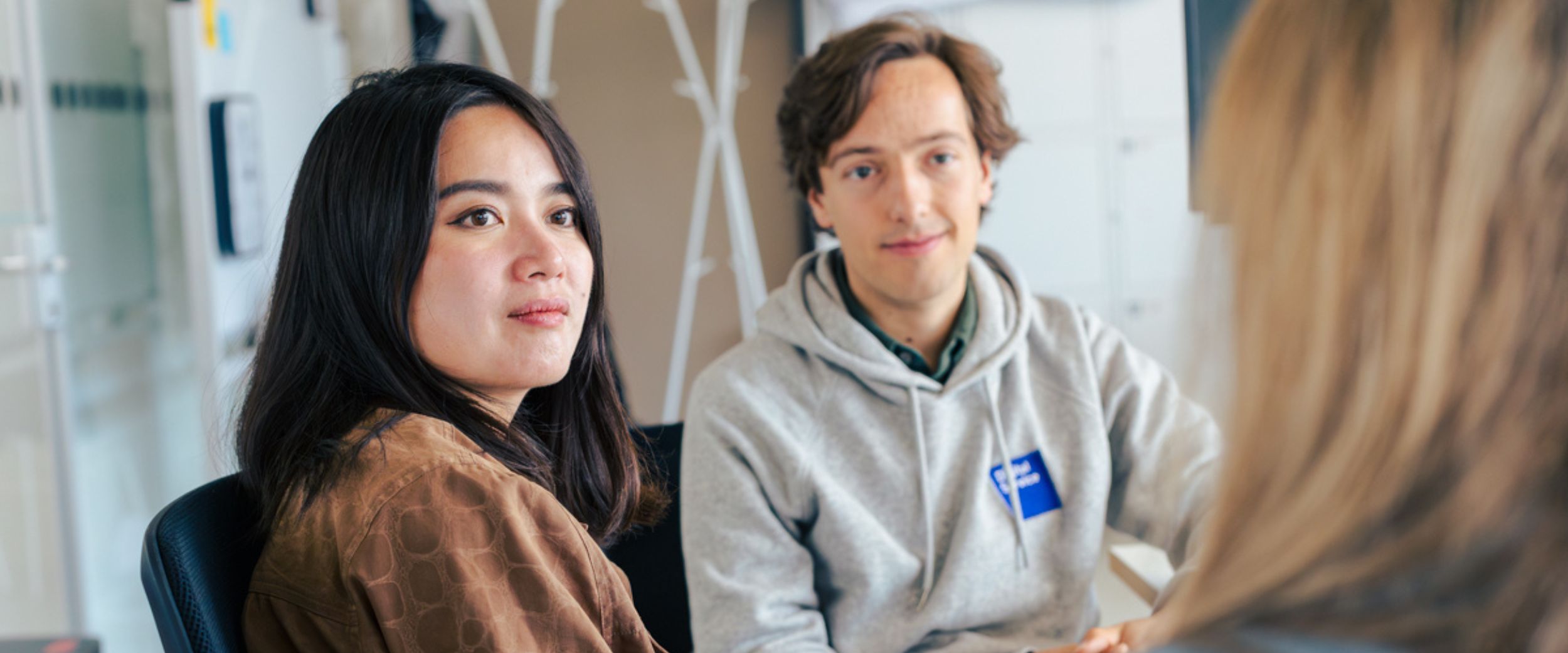Zwei Mitarbeitende des DigitalService sitzen in einem Büro und schauen eine dritte Person an, die nur von hinten zu sehen ist. Im Hintergrund steht ein Kleiderständer, Schränke und eine Pinnwand.
