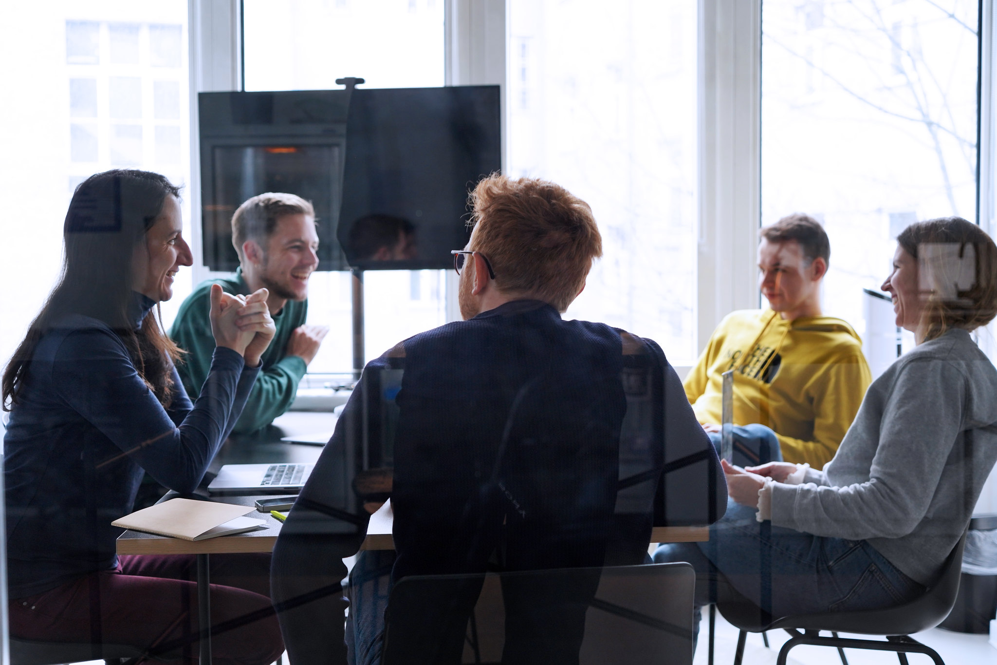 Five people sit at a table in a modern office and talk to each other – all smiling.
