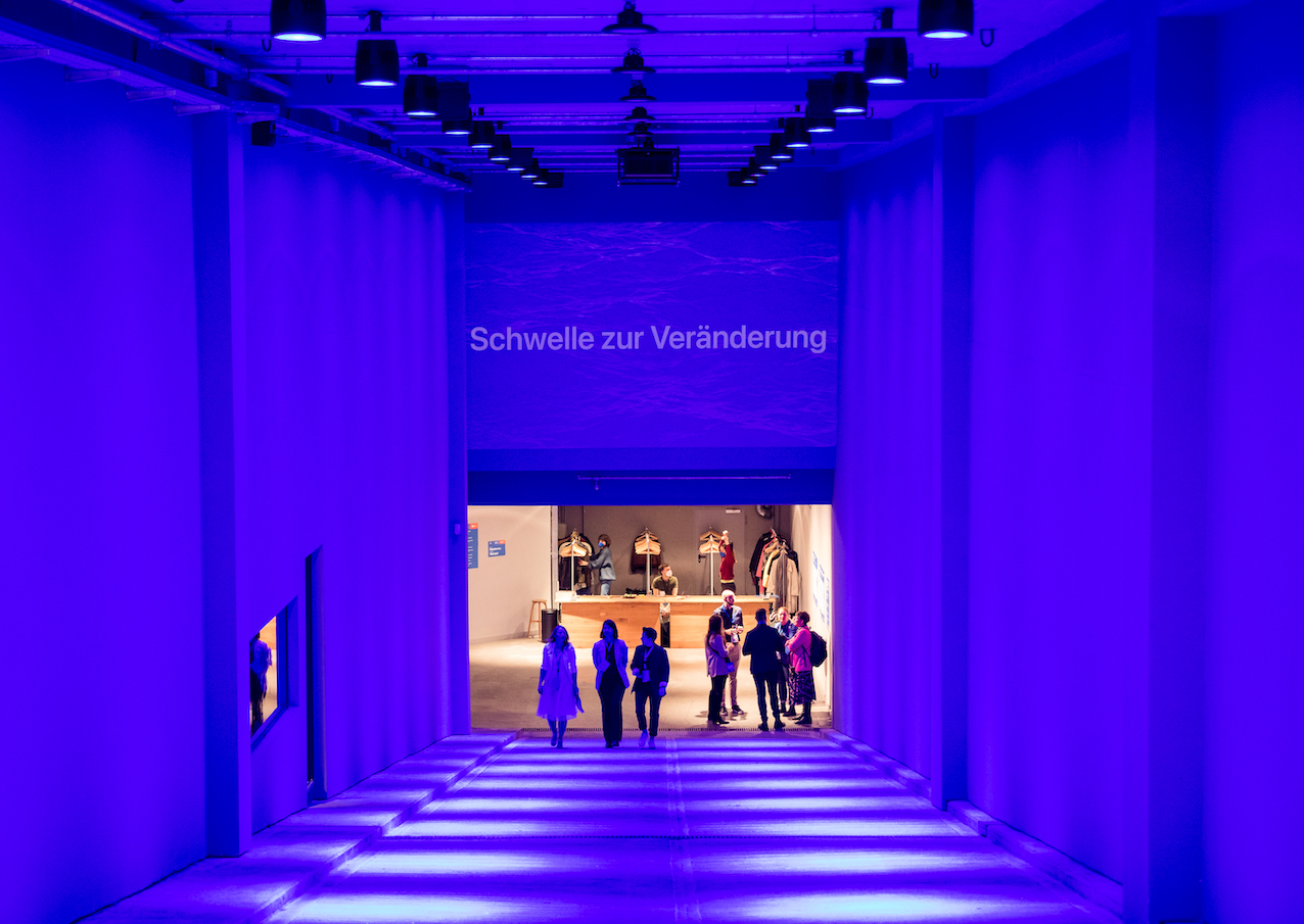 Eine Gruppe von Personen läuft in einem schräg abfallenden, tunnelartigen Zugang mit hohen Wänden nach unten in Richtung einer hell erleuchteten Garderobe; der Zugang ist in blaues Licht gehüllt; am oberen Rand des Zugangs steht „Schwelle zur Veränderung
