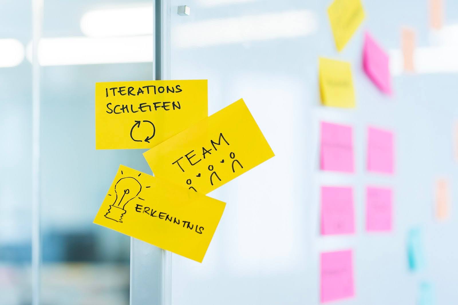 Auf einer Glaswand sind drei Zettel geklebt. Auf den Zetteln steht „Iterationsschleifen“, „Team“ und „Erkenntnis“.
