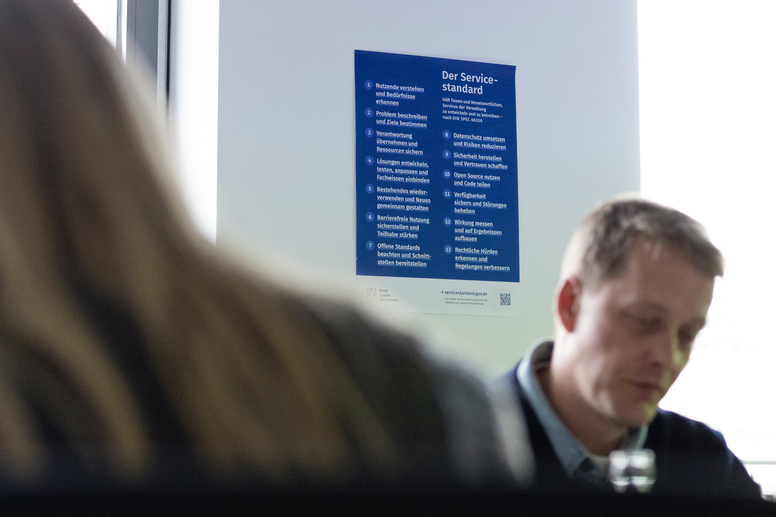 Ein Poster mit den 13 Punkten des Servicestandards an einer Wand während eines Workshops; 2 Personen sind unscharf im Vordergrund, sie sehen konzentriert aus