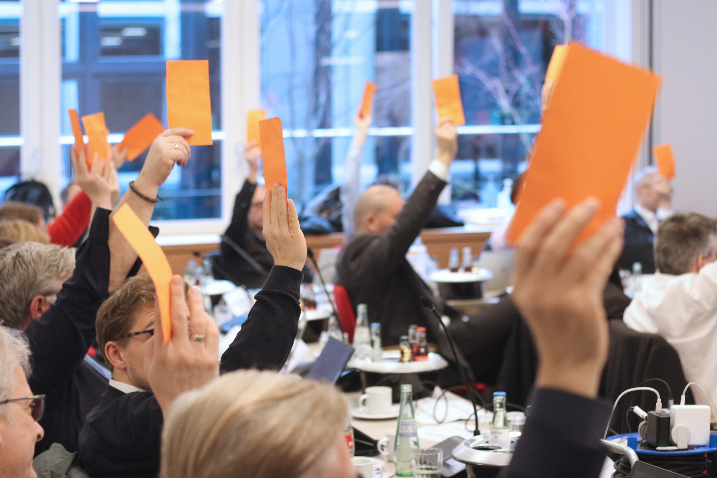 Eine Gruppe von Menschen in einem Sitzungssaal hebt orangefarbene Karten bei der Verabschiedung der DIN SPEC 66336. Die Personen sitzen an Tischen mit Getränken und Dokumenten, während sie aktiv an der Versammlung teilnehmen.
