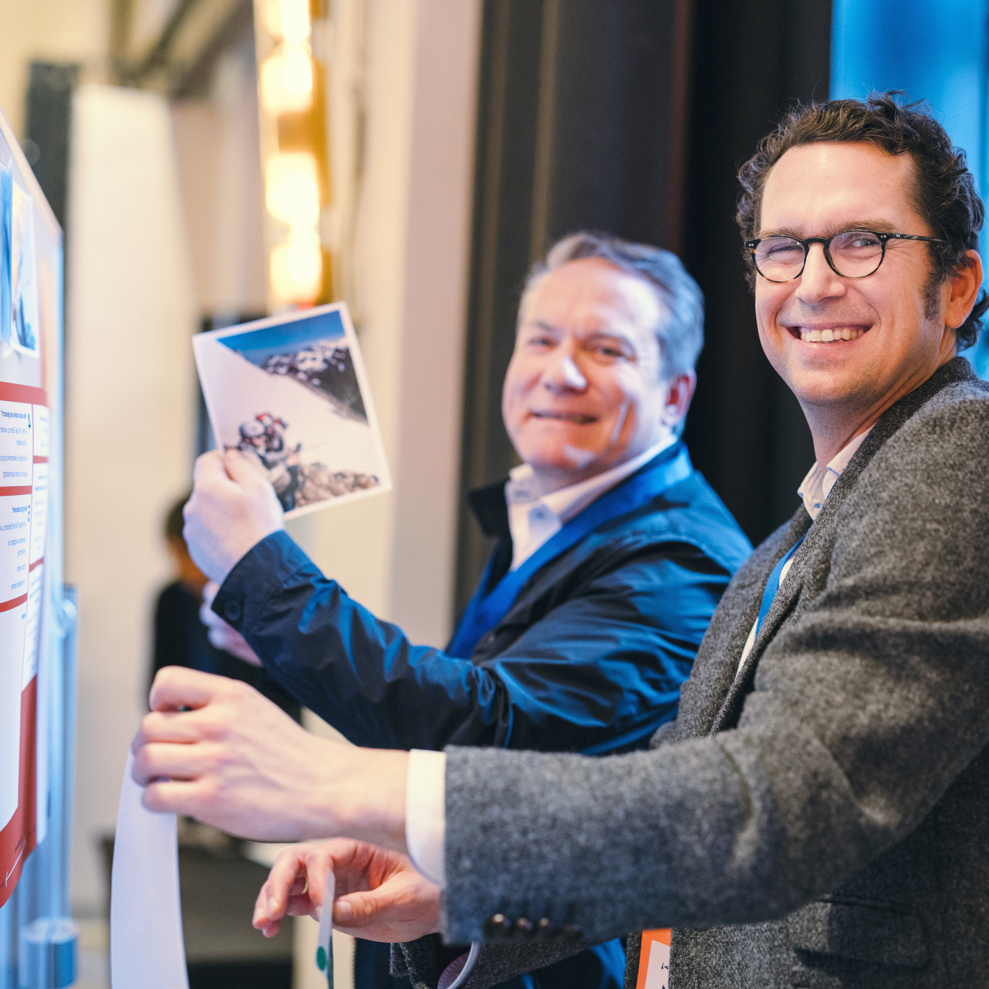 Zwei Personen schauen lachend in die Kamera, während sie Bilder und Notizen an eine Aufstellwand hängen