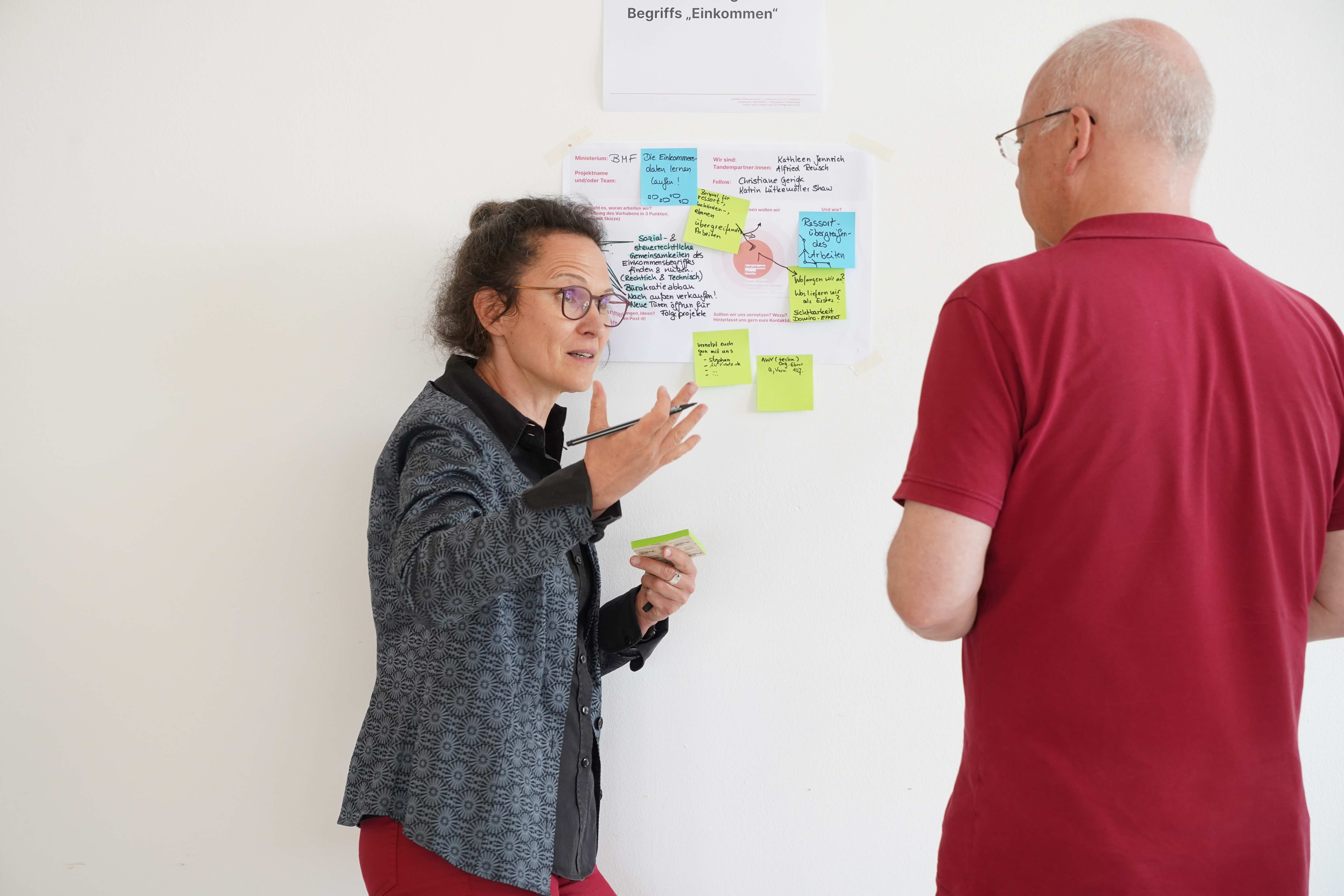 A woman stands in front of a wall with colorful sticky notes. She is gesticulating and talking to a man.
