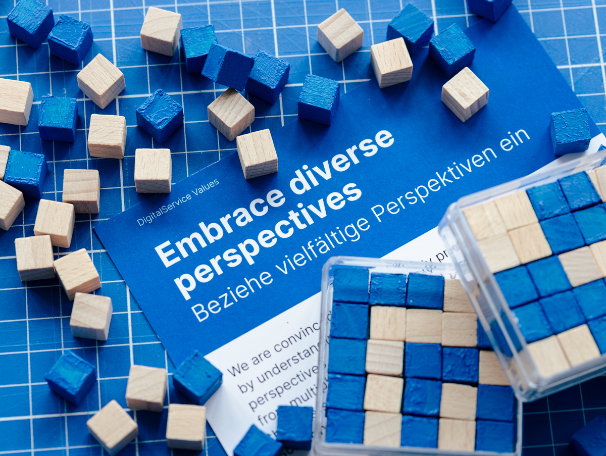 Holzwürfel, teils naturbelassen, teils blau bemalt, liegen verstreut auf kariertem Untergrund. Daneben ein Blatt mit dem Unternehmenswert „Embrace diverse perspectives / Beziehe vielfältige Perspektiven ein“ und Kästen mit Würfeln in Mustern.