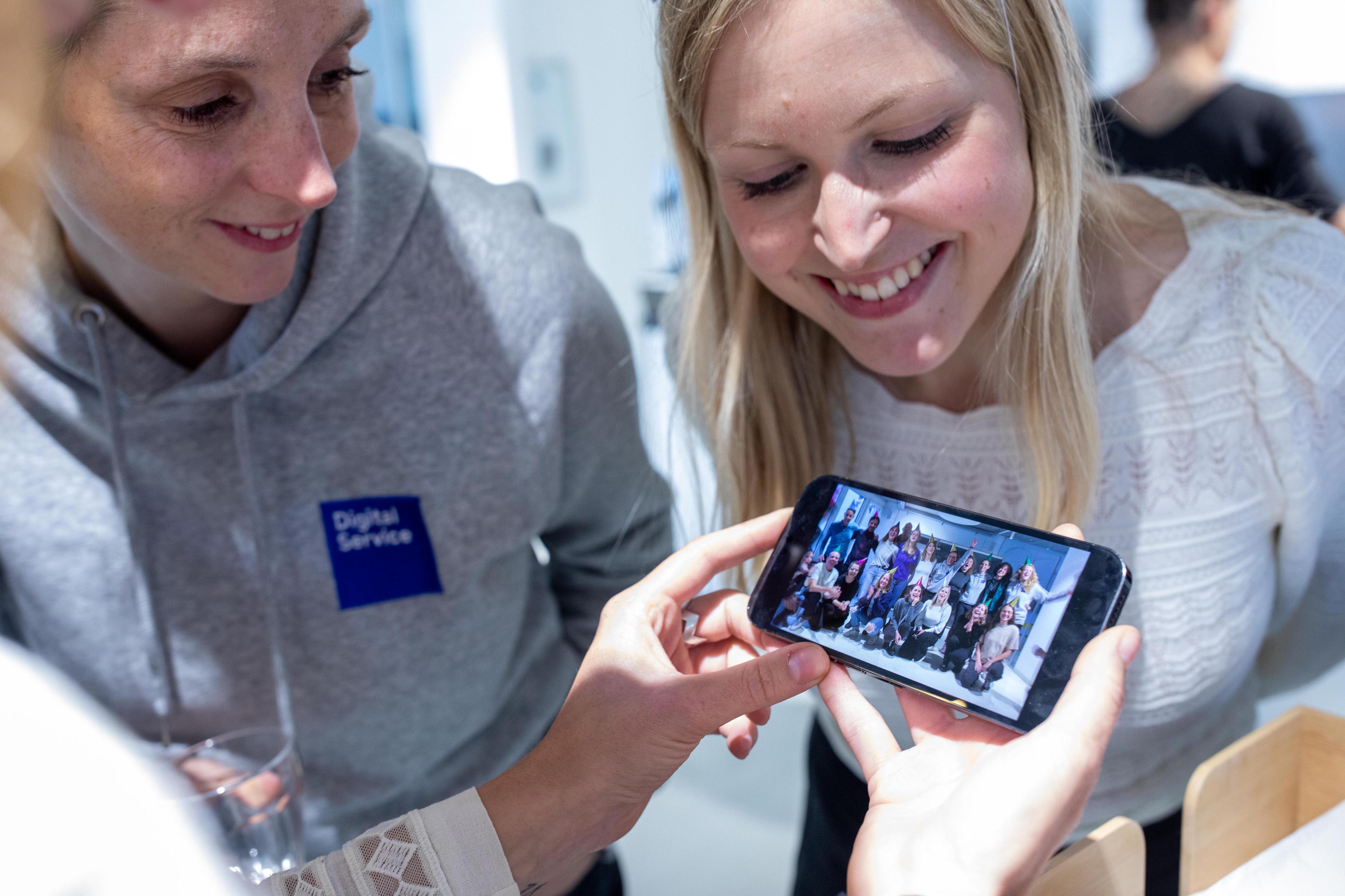 Zwei lachende Personen schauen auf ein Smartphone, das ein Gruppenfoto zeigt. Eine der Personen trägt einen grauen Hoodie mit dem Logo „DigitalService“.