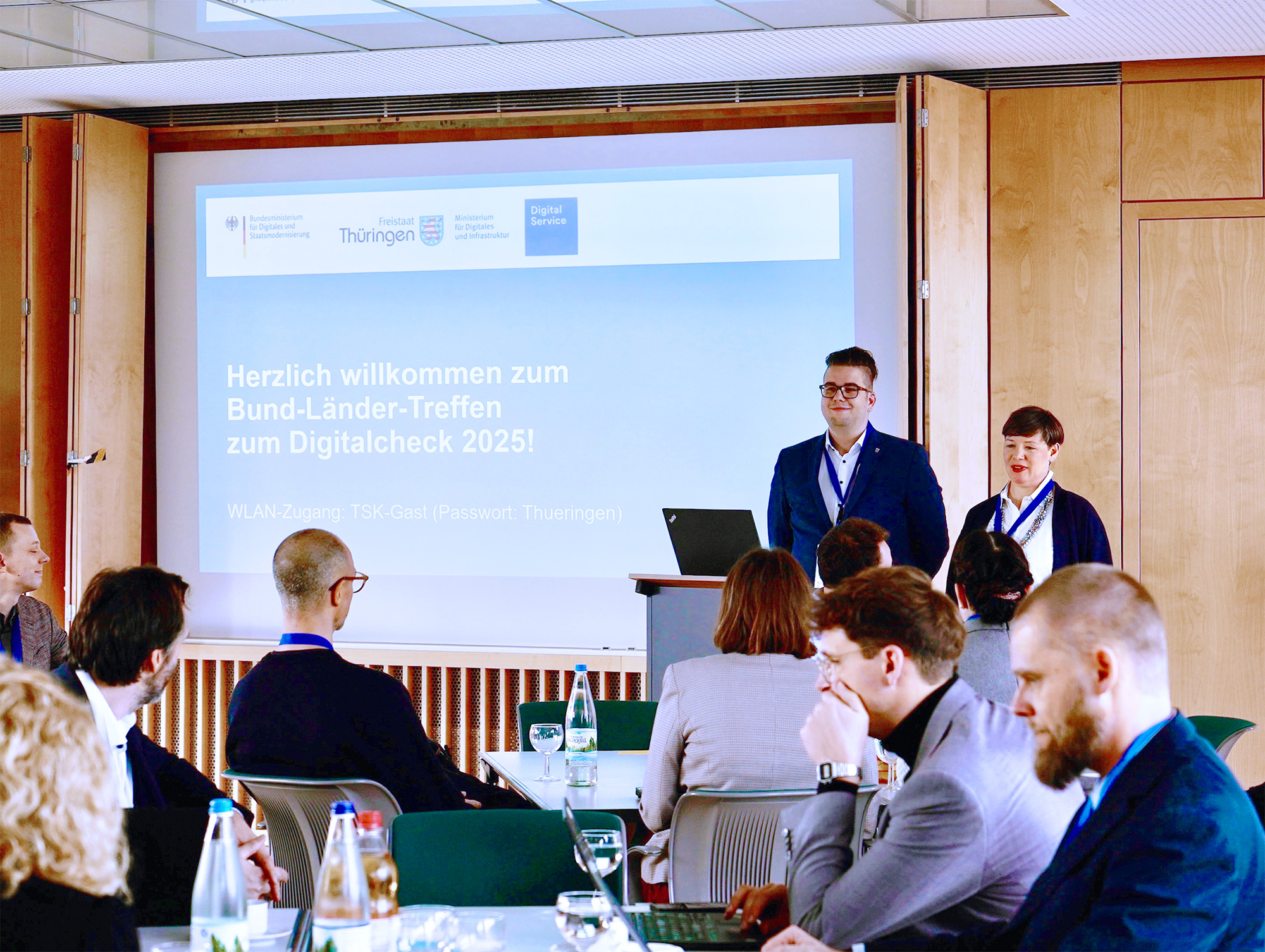 Zwei Personen stehen in einem Raum und präsentieren etwas, auf einer Leinwand hinter ihnen steht: „Herzlich willkommen zum Bund-Länder-Treffen zum Digitalcheck 2025!“. Im Vordergrund sitzen weitere Personen und hören zu.