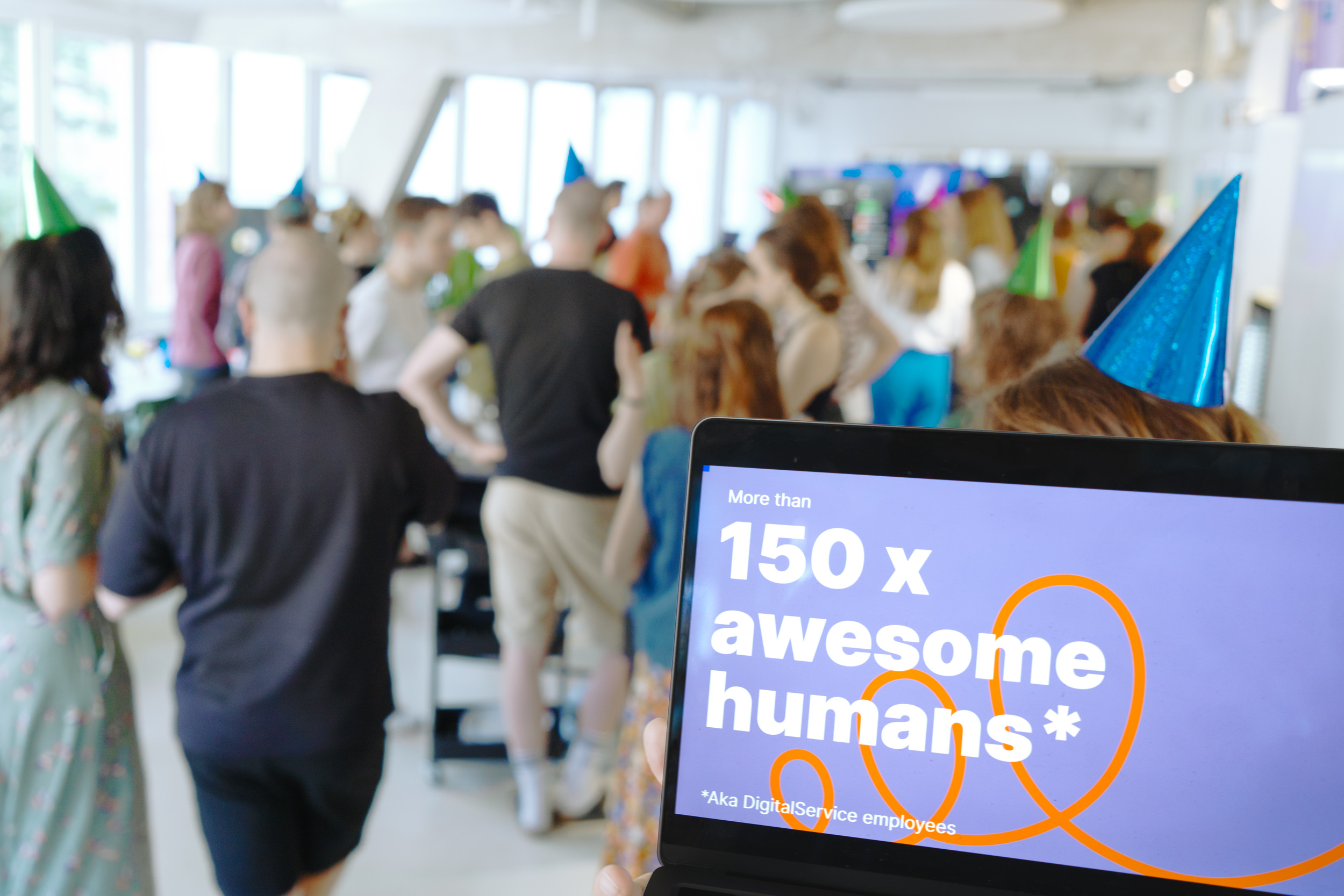 Im Vordergrund ist auf einem Laptopbildschirm zu lesen, dass mehr als 150 Mitarbeitende nun beim DigitalService arbeiten; im Hintergrund sind unscharf zahlreiche Mitarbeitende zu erkennen, die gemeinsam feiern