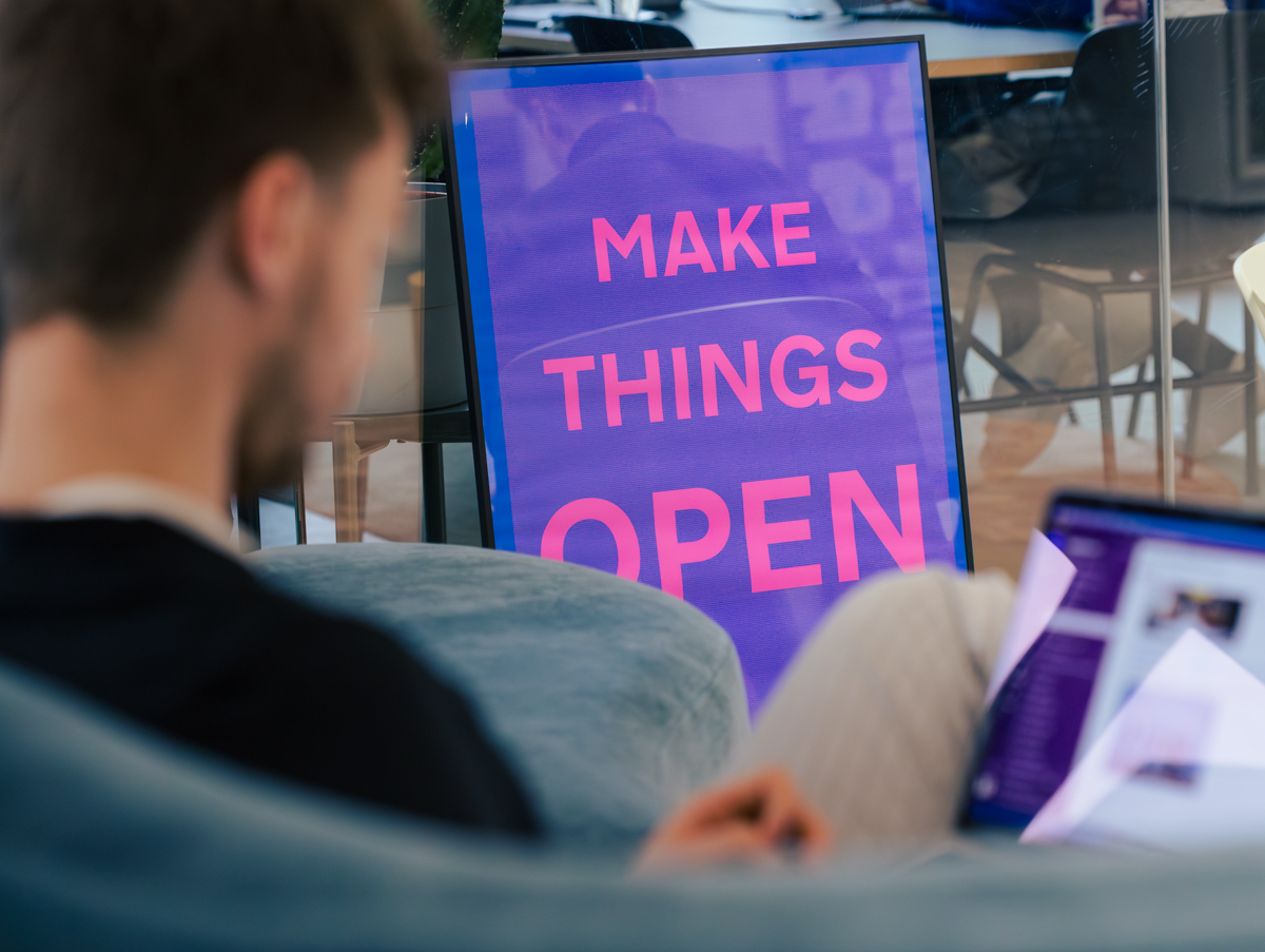 Ein Poster mit dem Slogan „Make Things Open“ steht an einer Glaswand im DigitalService Büro; im Vordergrund sitz ein DigitalService Mitarbeitender, der schemenhaft zu erkennen ist